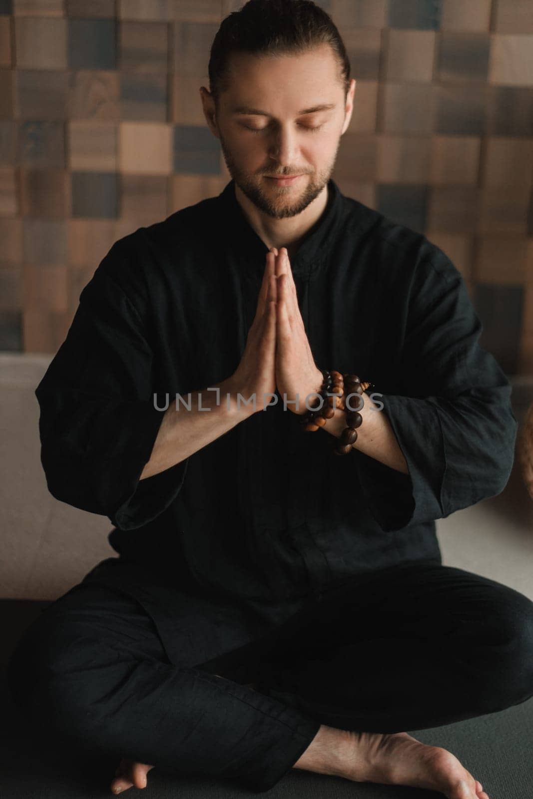 Portrait of a young man in a black kimano sitting in a lotus position on a gym mat in the interior.