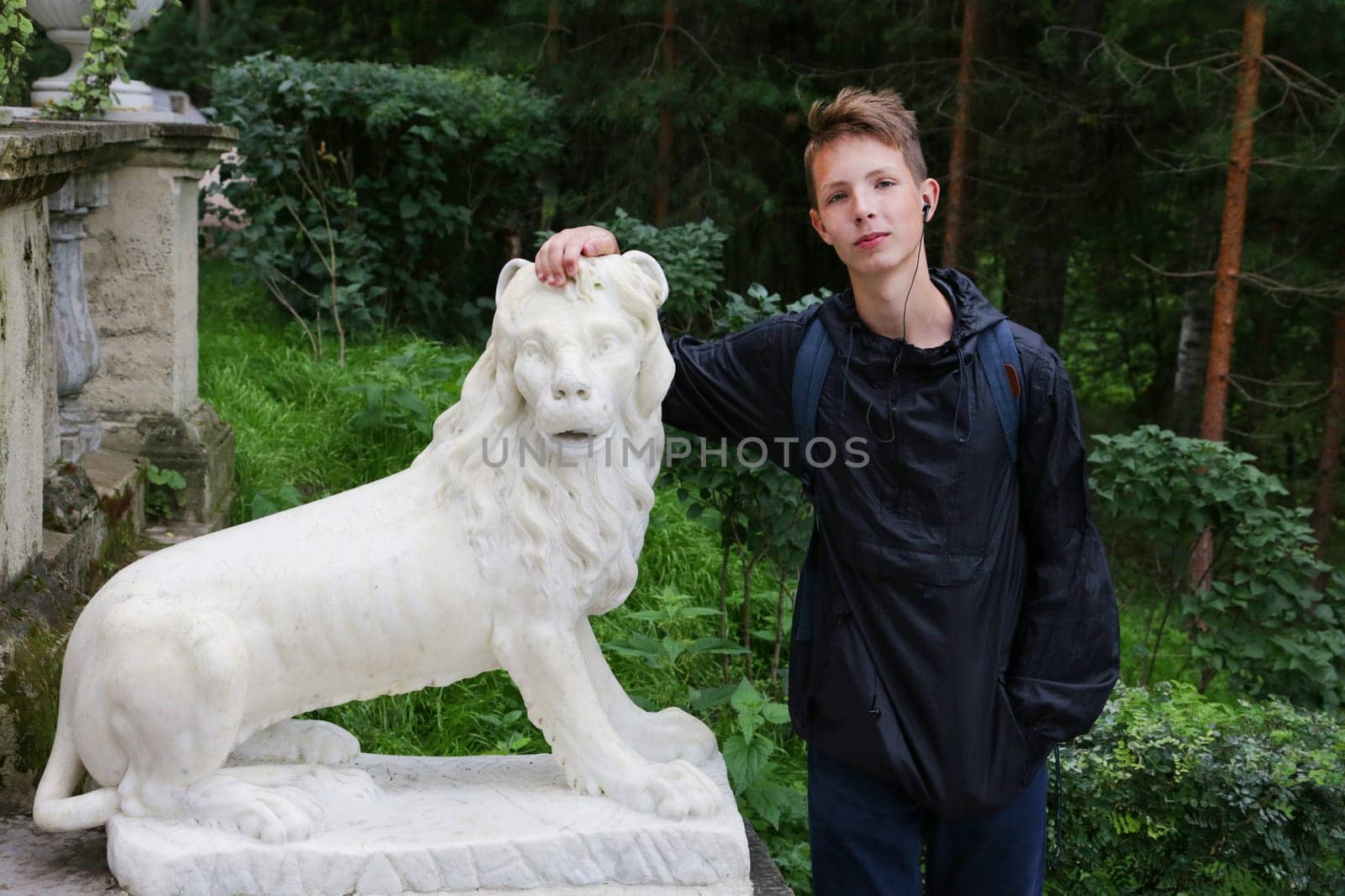 A handsome and cheerful boy, age 14-17, stands near a white stone lion statue against a green backdrop. The boy in blue clothes is leaning his hand on the lion statue.