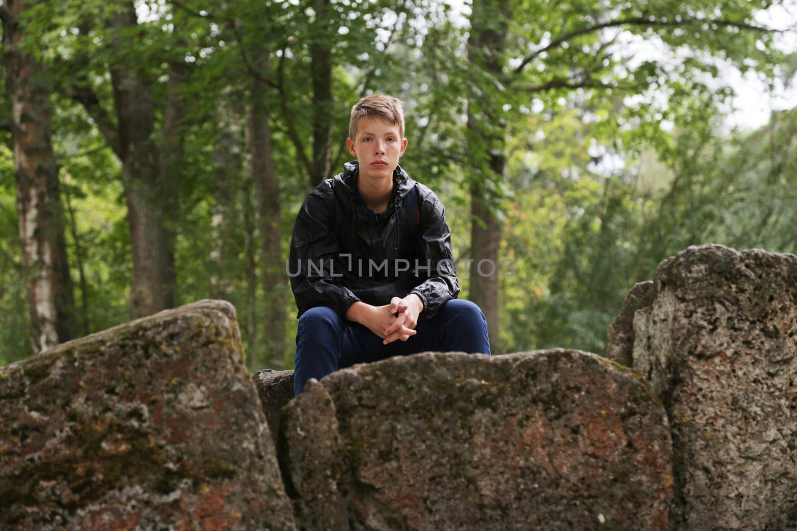 A handsome young man in a blue hiking jacket from the rain sits on a large stone in the background of a forest with trees. Boy tourist on vacation.
