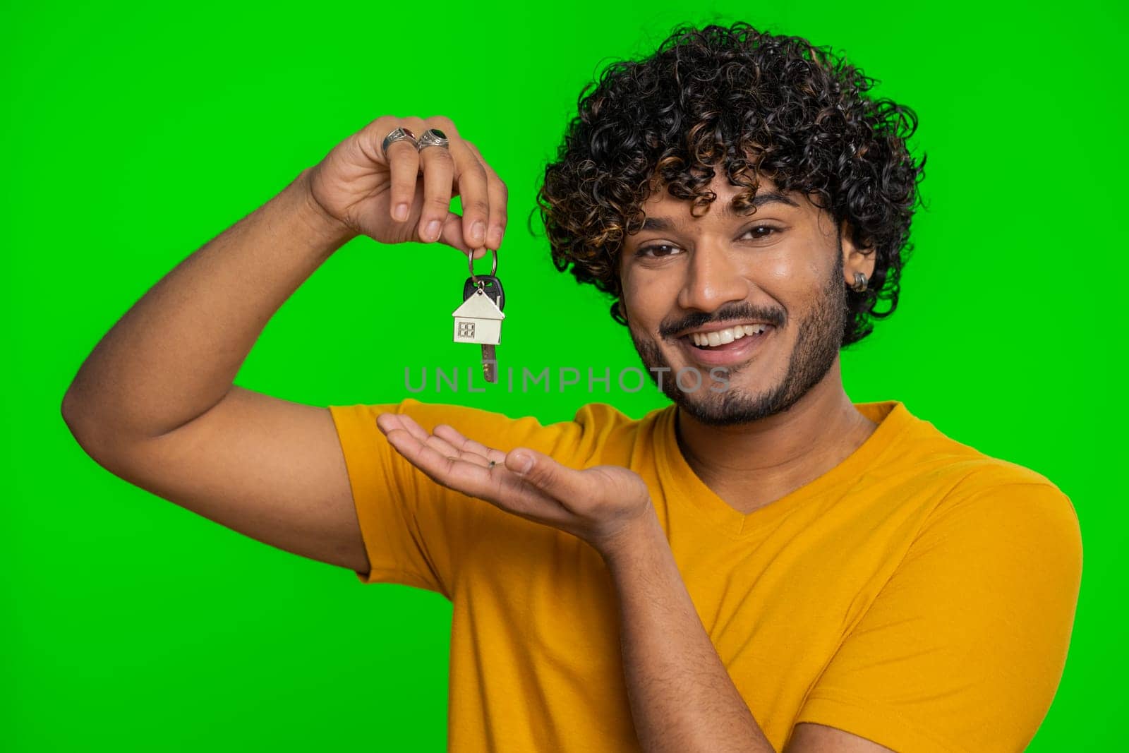 Young indian man real estate agent lifting hand showing the keys of new home house apartment, buying or renting property, mortgage loan. Bearded hindu guy isolated on chroma key background, indoors