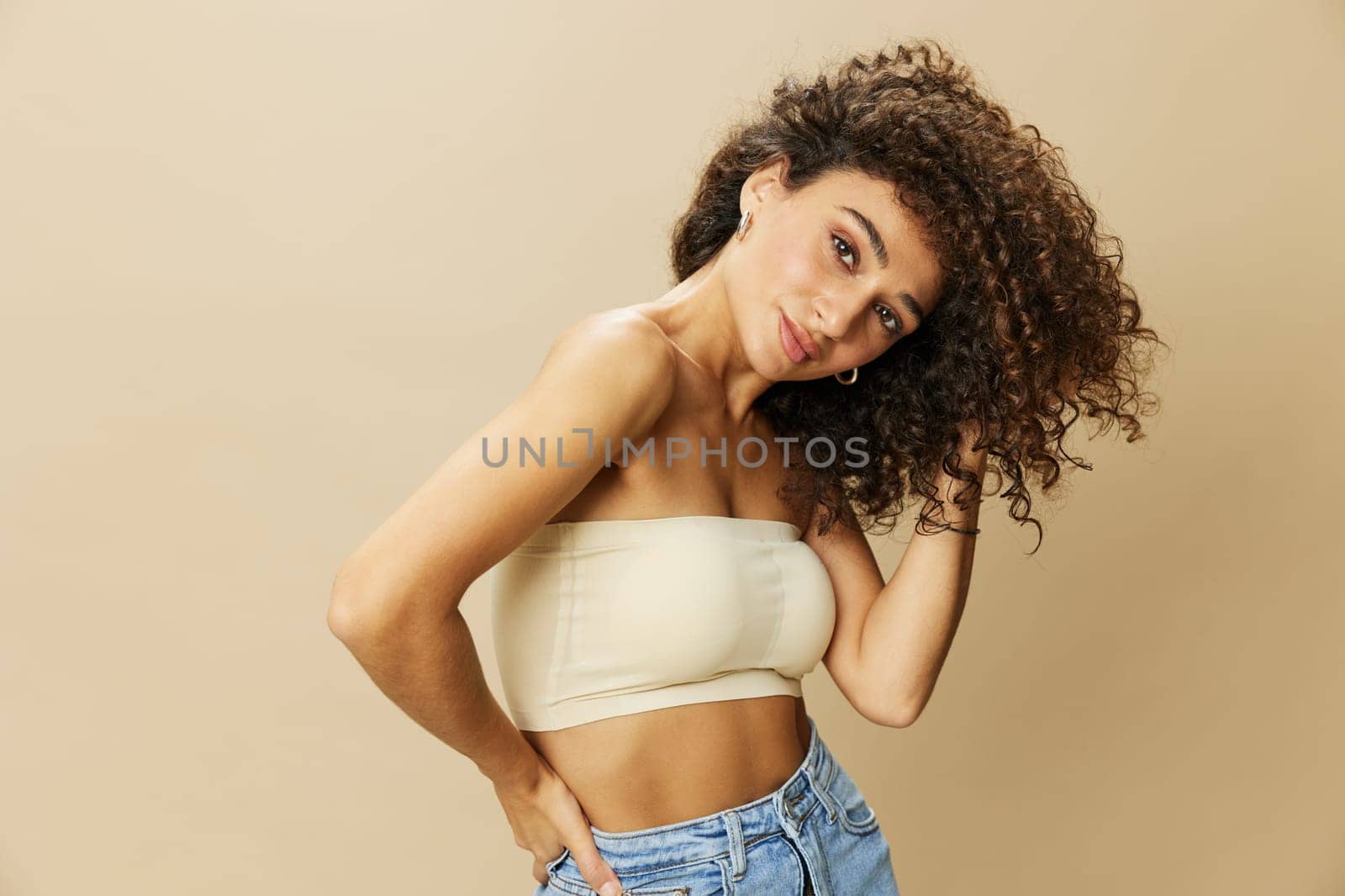 Woman applies cream and balm to her curly hair, the concept of protection and care, a healthy look, a smile with teeth on a beige background by SHOTPRIME