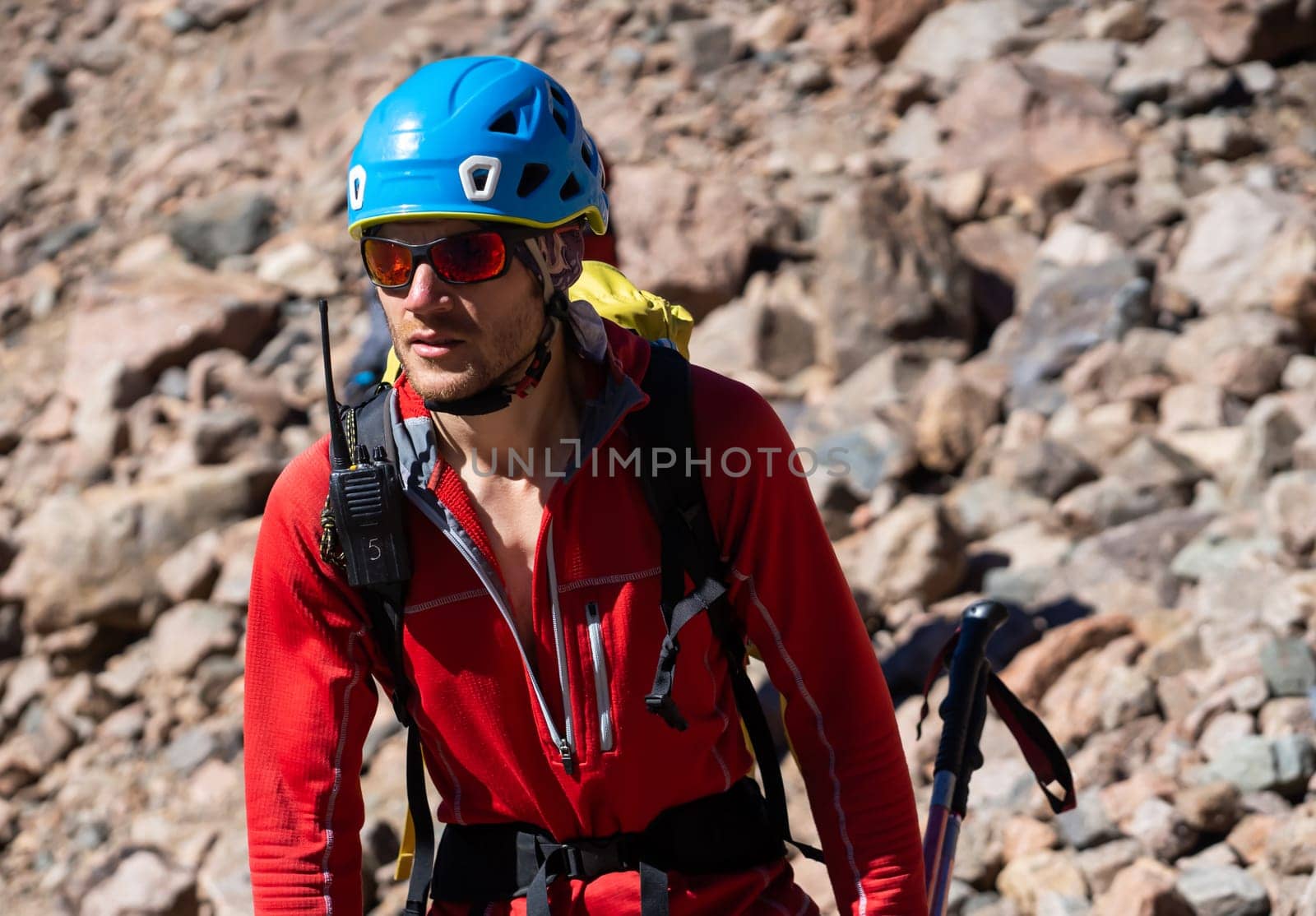 A young man in sunglasses, helmet and a red thermal jacket, with a walkie-talkie walks in the mountains alone. The traveler climbs the rocks, stones, goes in for mountaineering.