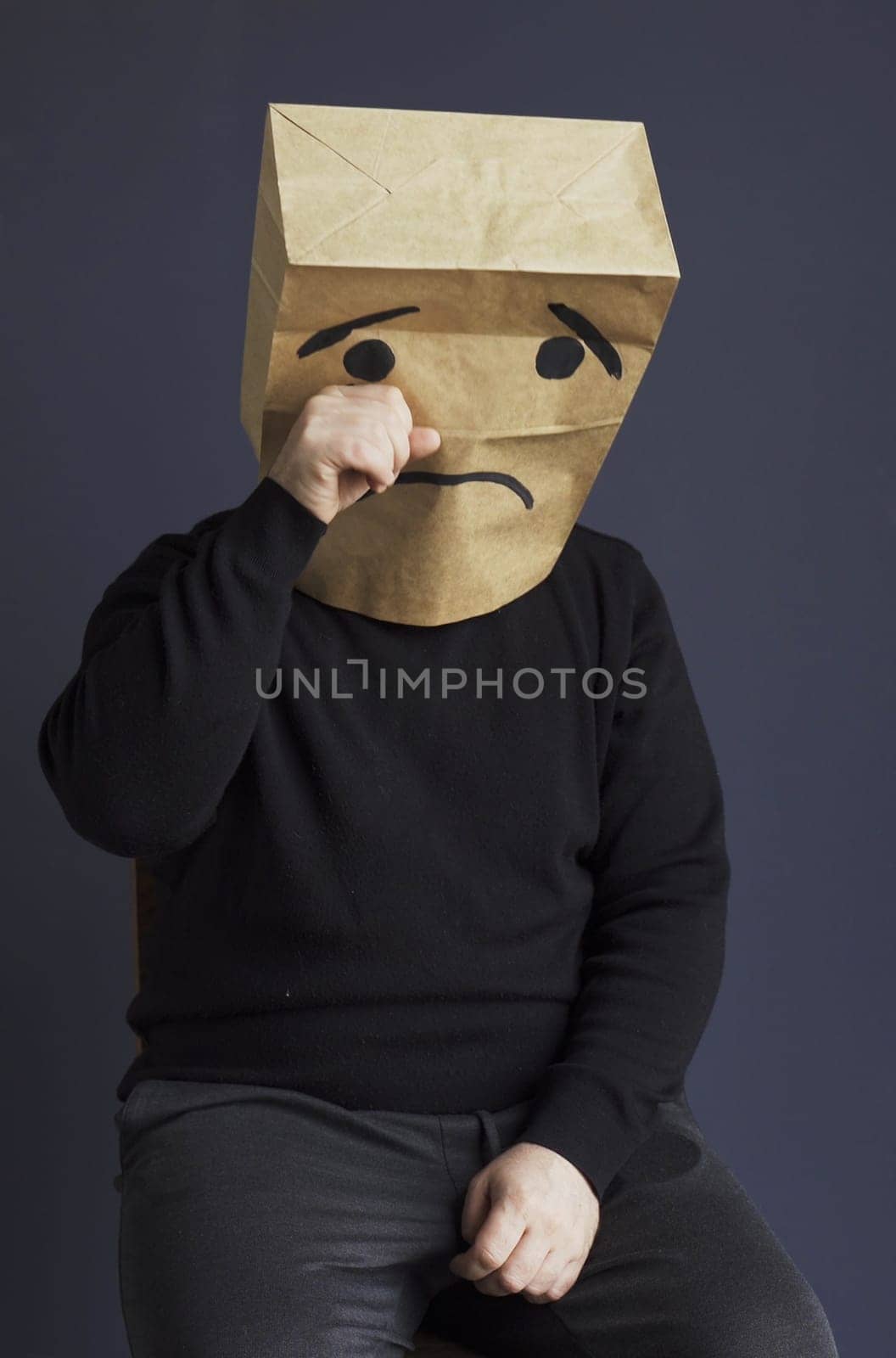 A sad man in a black turtleneck with a bag on his head, with a drawn crying emoticon, cries and wipes away a tear. Emotions and gestures. Vertical frame.