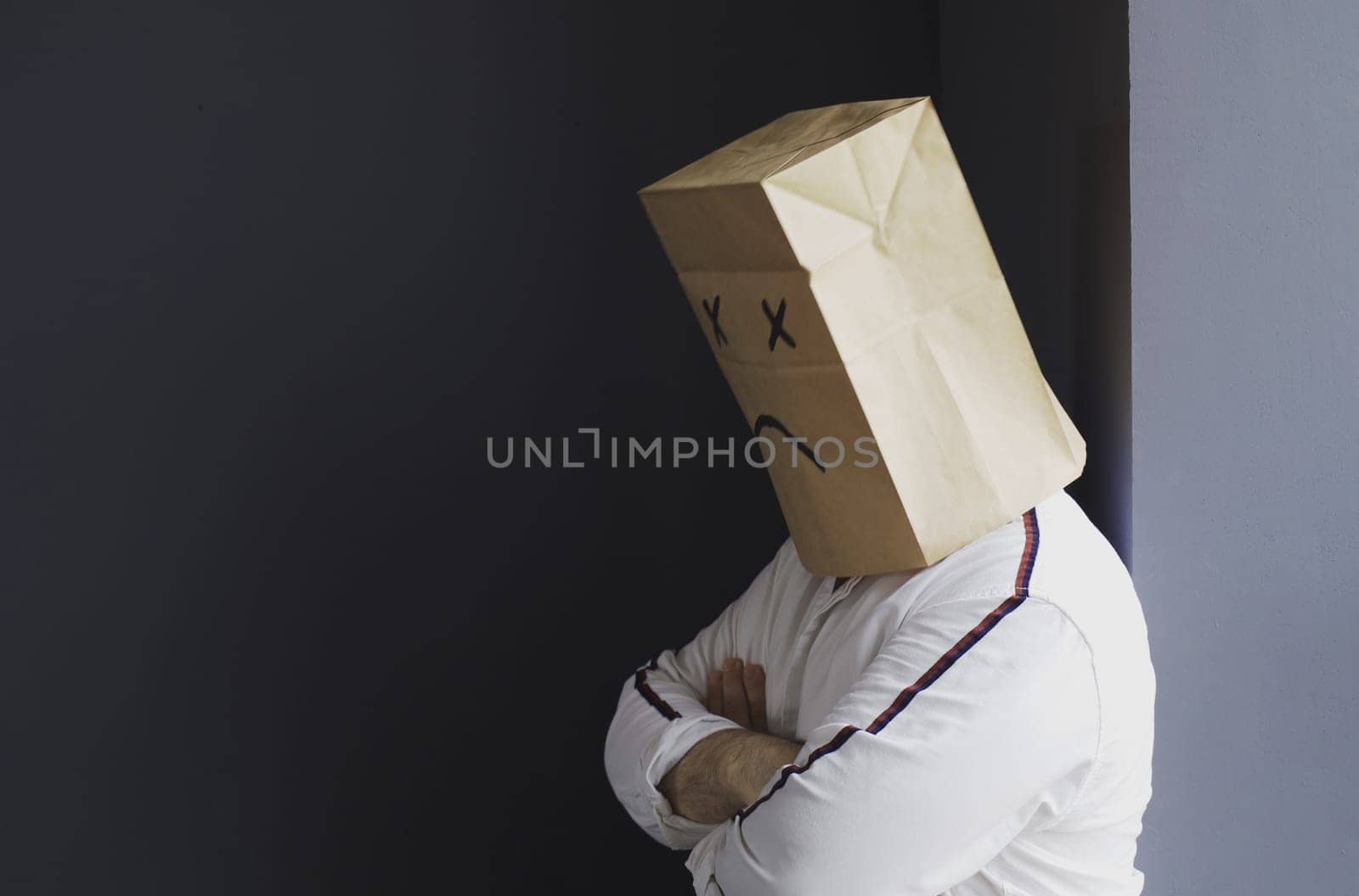 A sad man in a white shirt with a bag on his head, with a sad smiley face drawn, stands against the wall and cries.