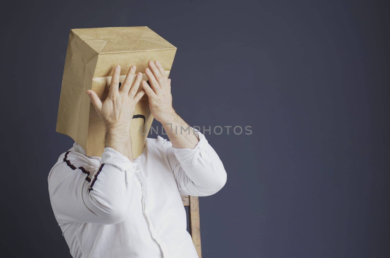 A sad man in a white shirt with a bag on his head, with a drawn sad emoticon is afraid, closes his eyes with his hands. Emotions and gestures.