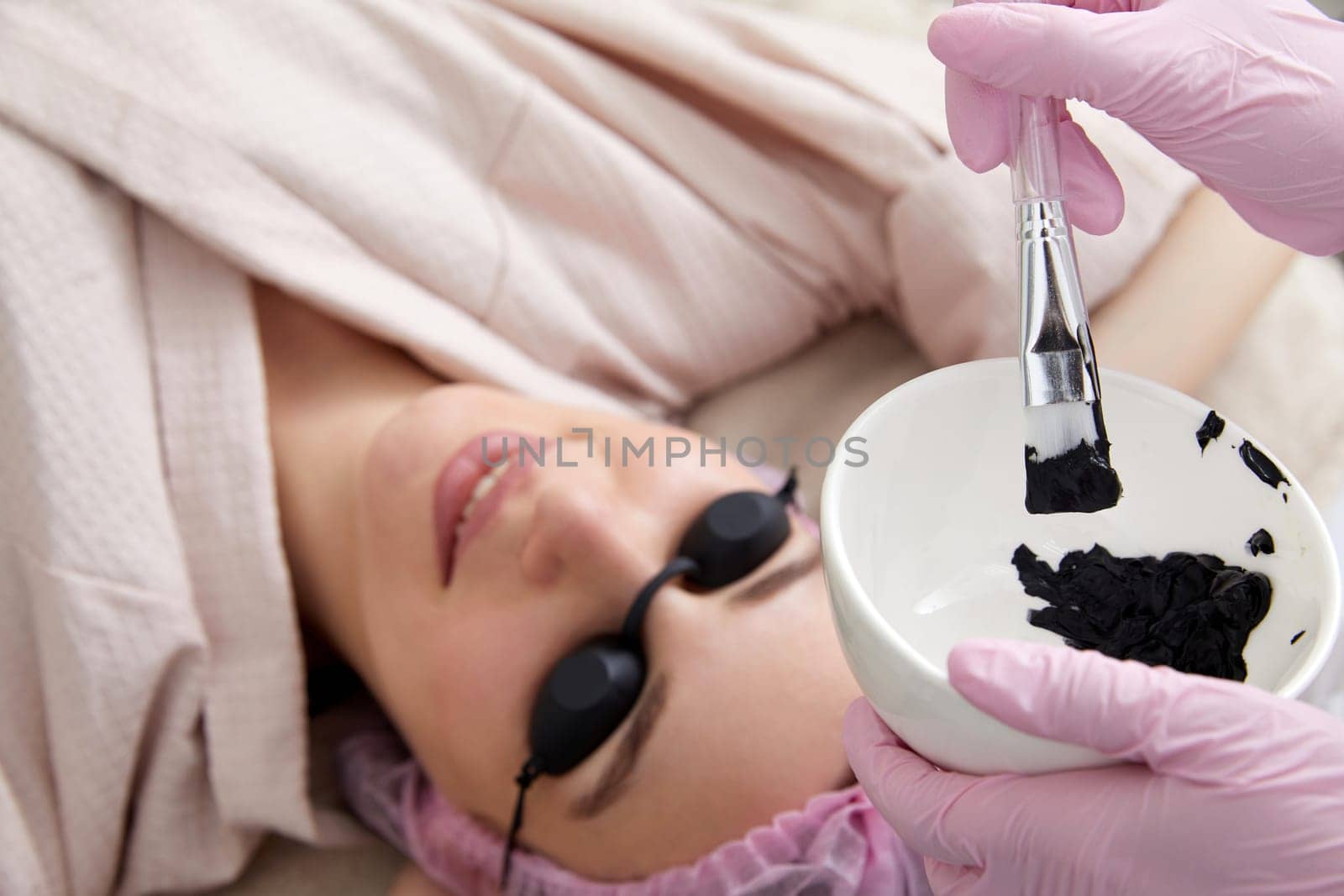 Carbon peeling in beauty salon. Cosmetologist applying black mask on the face of beautiful woman for carbon peeling
