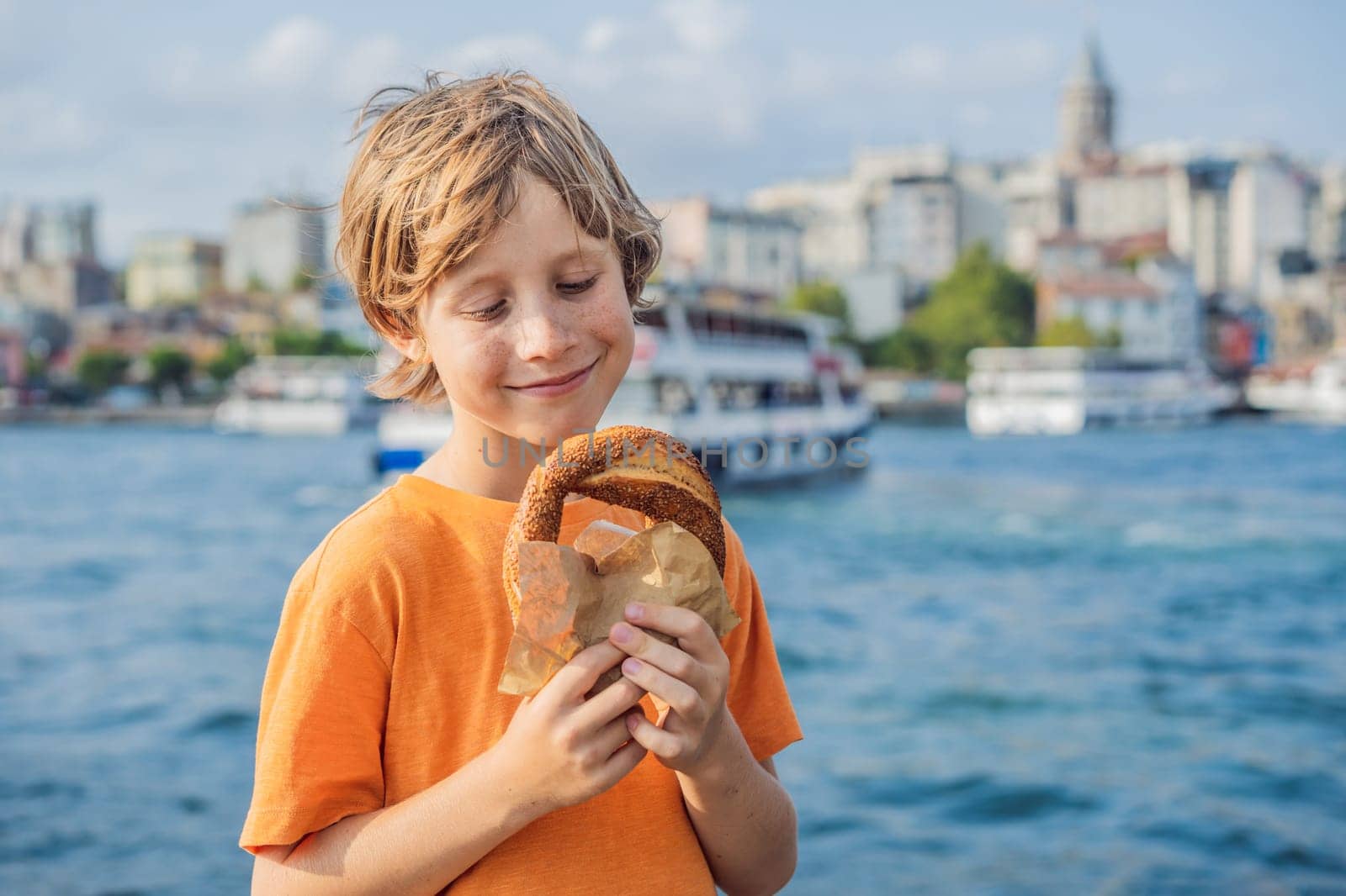 Boy in Istanbul having breakfast with Simit and a glass of Turkish tea. Glass of Turkish tea and bagel Simit against golden horn bay and the Galata Tower in Istanbul, Turkey. Turkiye. Traveling with kids concept.