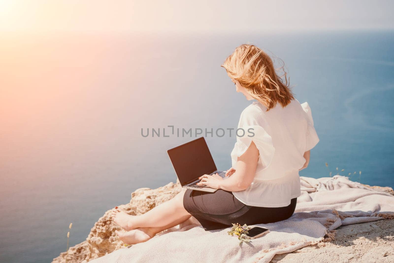 Successful business woman in yellow hat working on laptop by the sea. Pretty lady typing on computer at summer day outdoors. Freelance, travel and holidays concept.