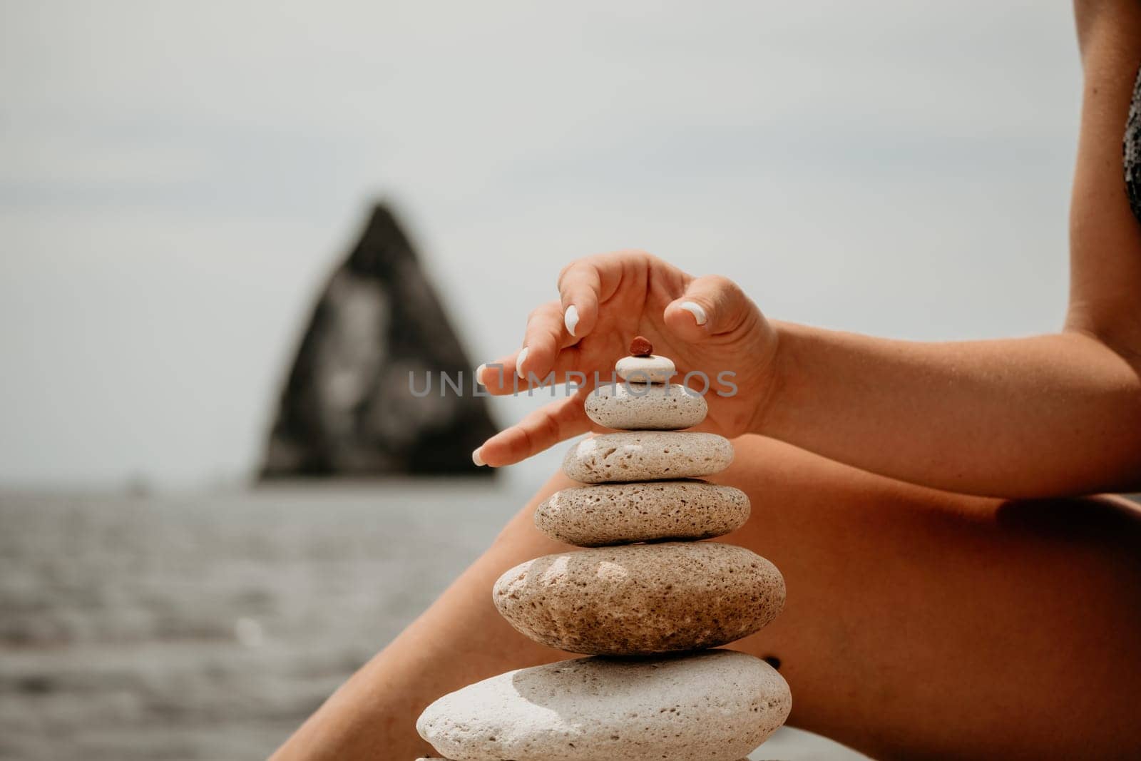 Woman bilds stones pyramid on the seashore on a sunny day on the blue sea background. Happy holidays. Pebble beach, calm sea, travel destination. Concept of happy vacation on the sea, meditation, spa