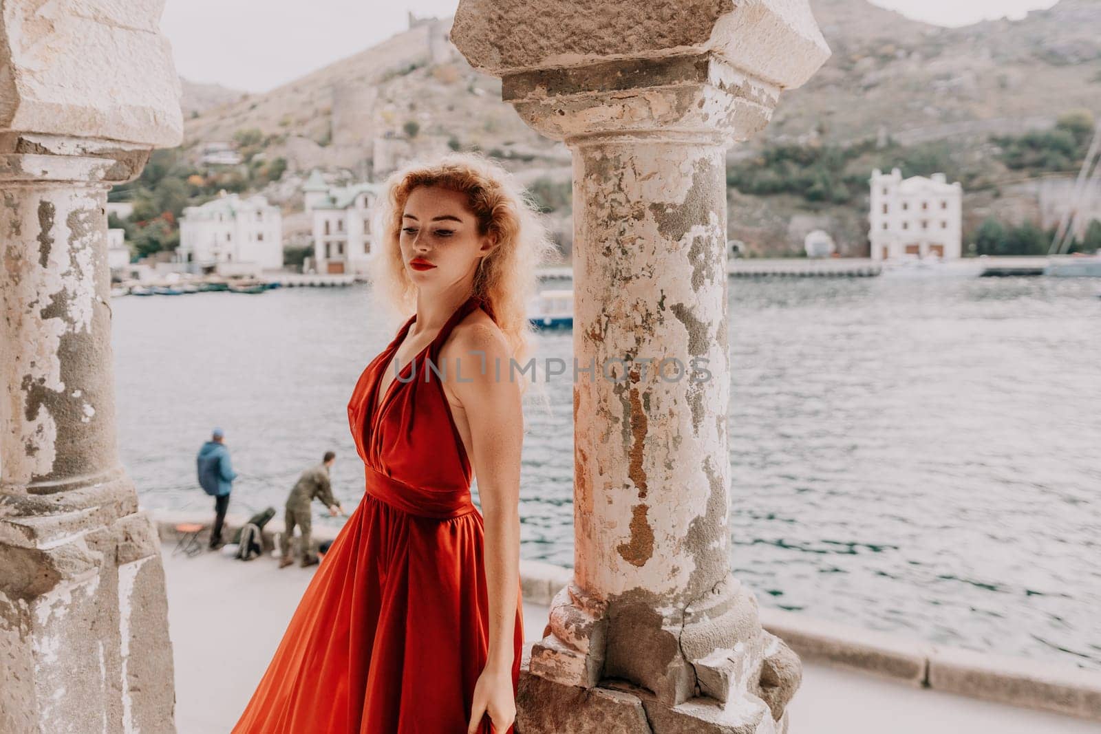 Close up shot of beautiful young caucasian woman with curly blond hair and freckles looking at camera and smiling. Cute woman portrait in a pink long dress posing on a volcanic rock high above the sea