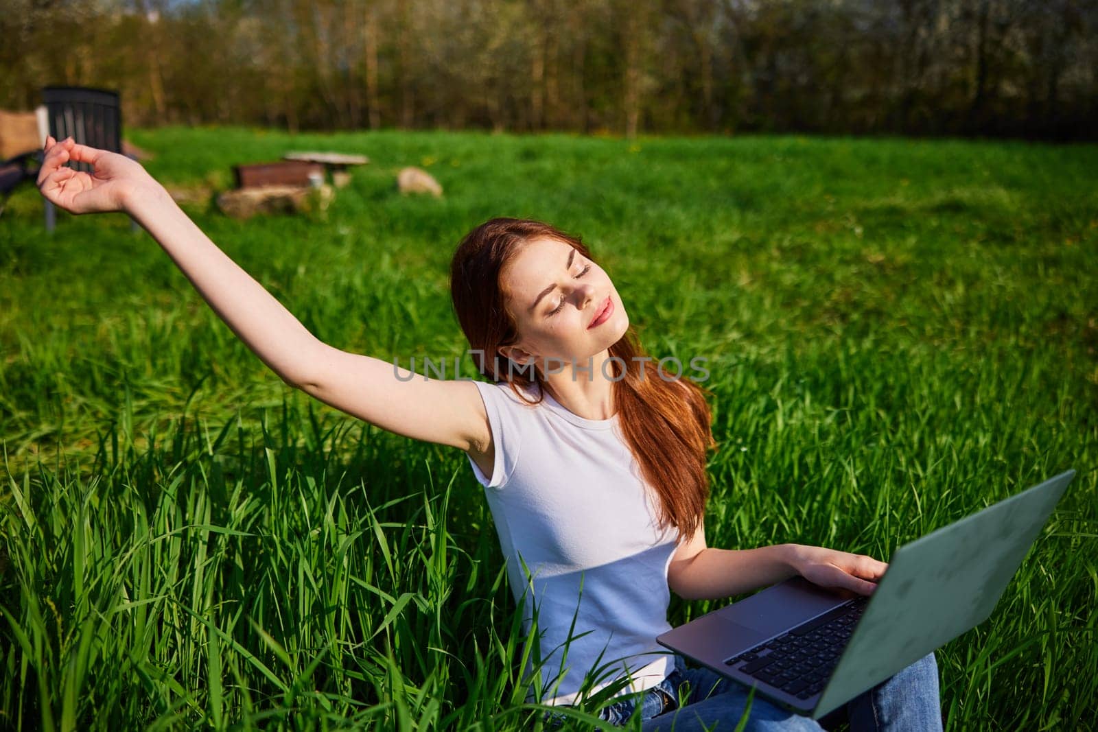 joyful woman works sitting in high grass behind a laptop raising her hand up. High quality photo