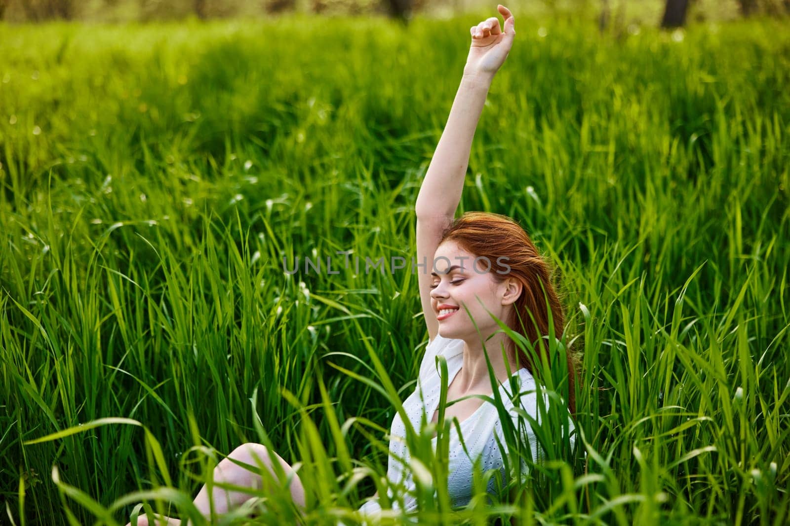 a cute woman in the summer high grass sits in a light dress happily raising her hand up. High quality photo