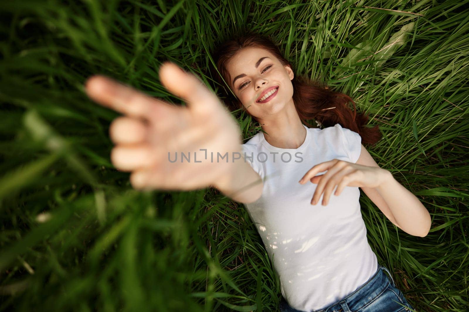 A happy girl in a white t-shirt stretches her arms up and lies on the green grass by Vichizh