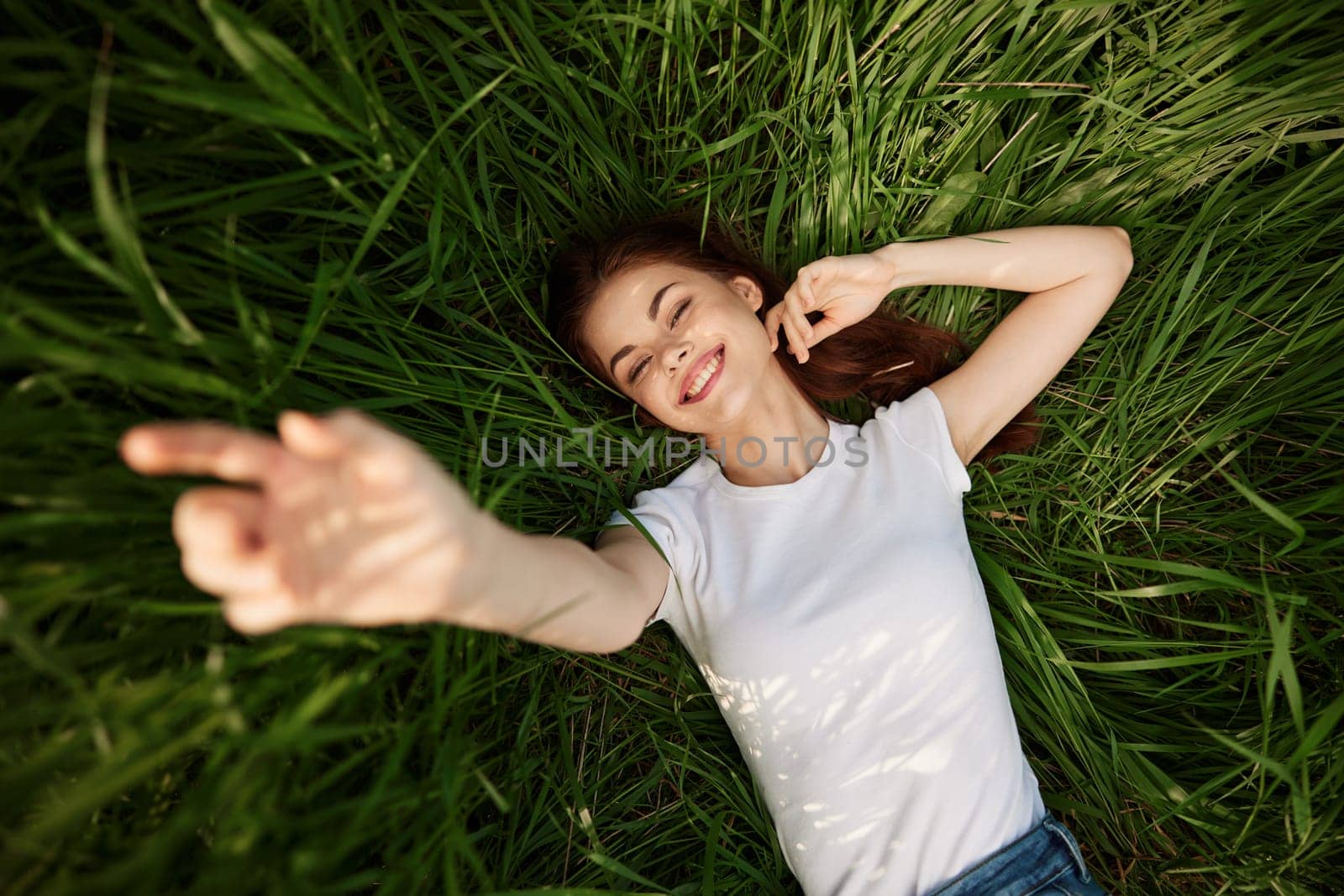 A happy girl in a white t-shirt stretches her arms up and lies on the green grass. High quality photo