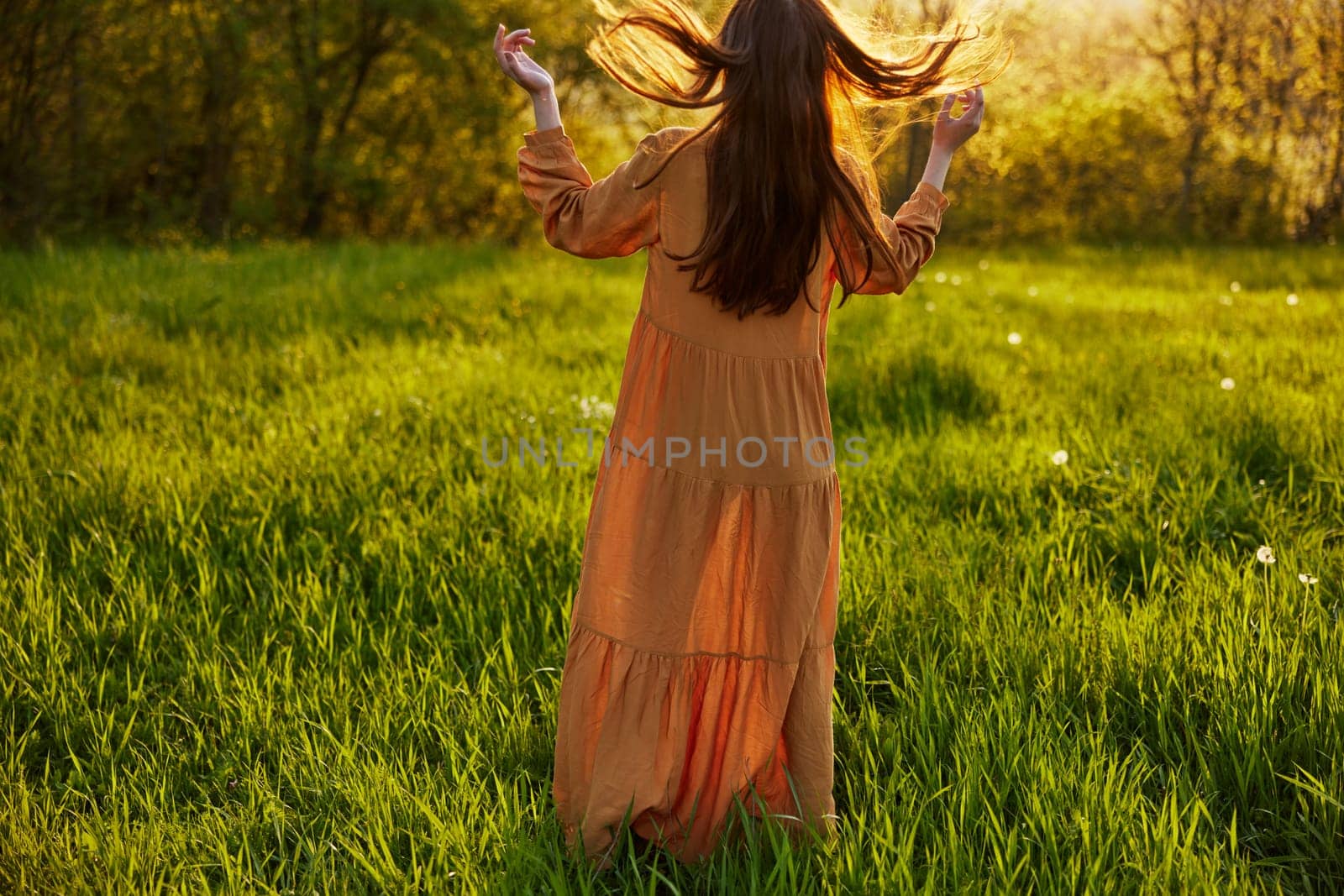 a slender woman stands in a long orange dress with her back to the camera illuminated by the sunset rays and tosses her long hair. High quality photo