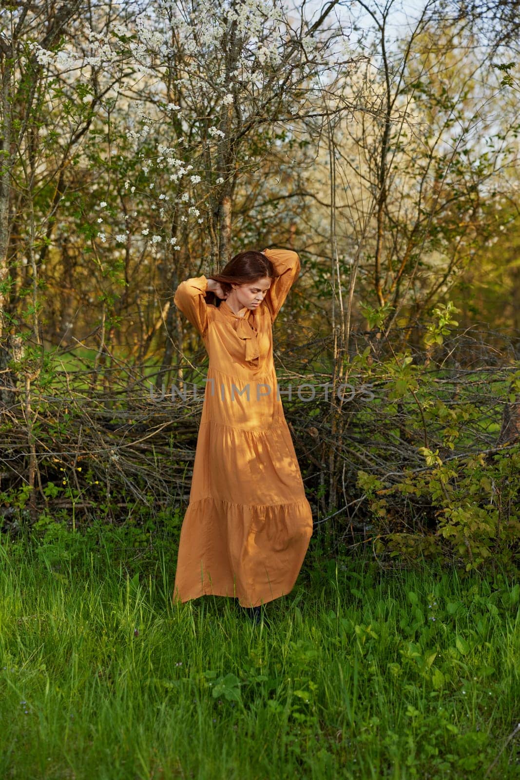 an elegant, sophisticated woman poses relaxed standing near a wicker fence at the dacha in a long orange dress holding her hair with her hands. High quality photo