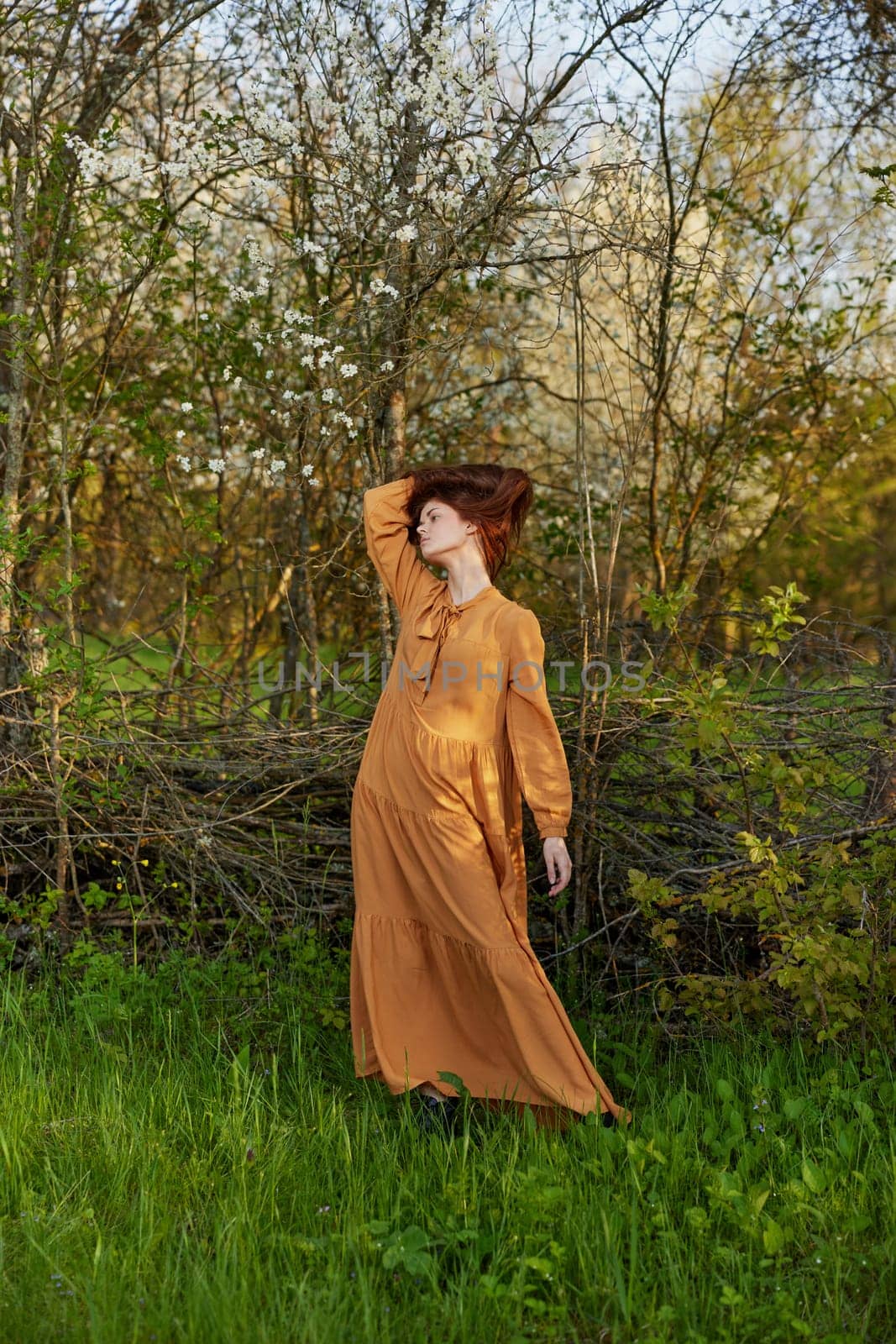 an elegant, sophisticated woman poses relaxed standing near a wicker fence at the dacha in a long orange dress holding her hair with her hands. High quality photo