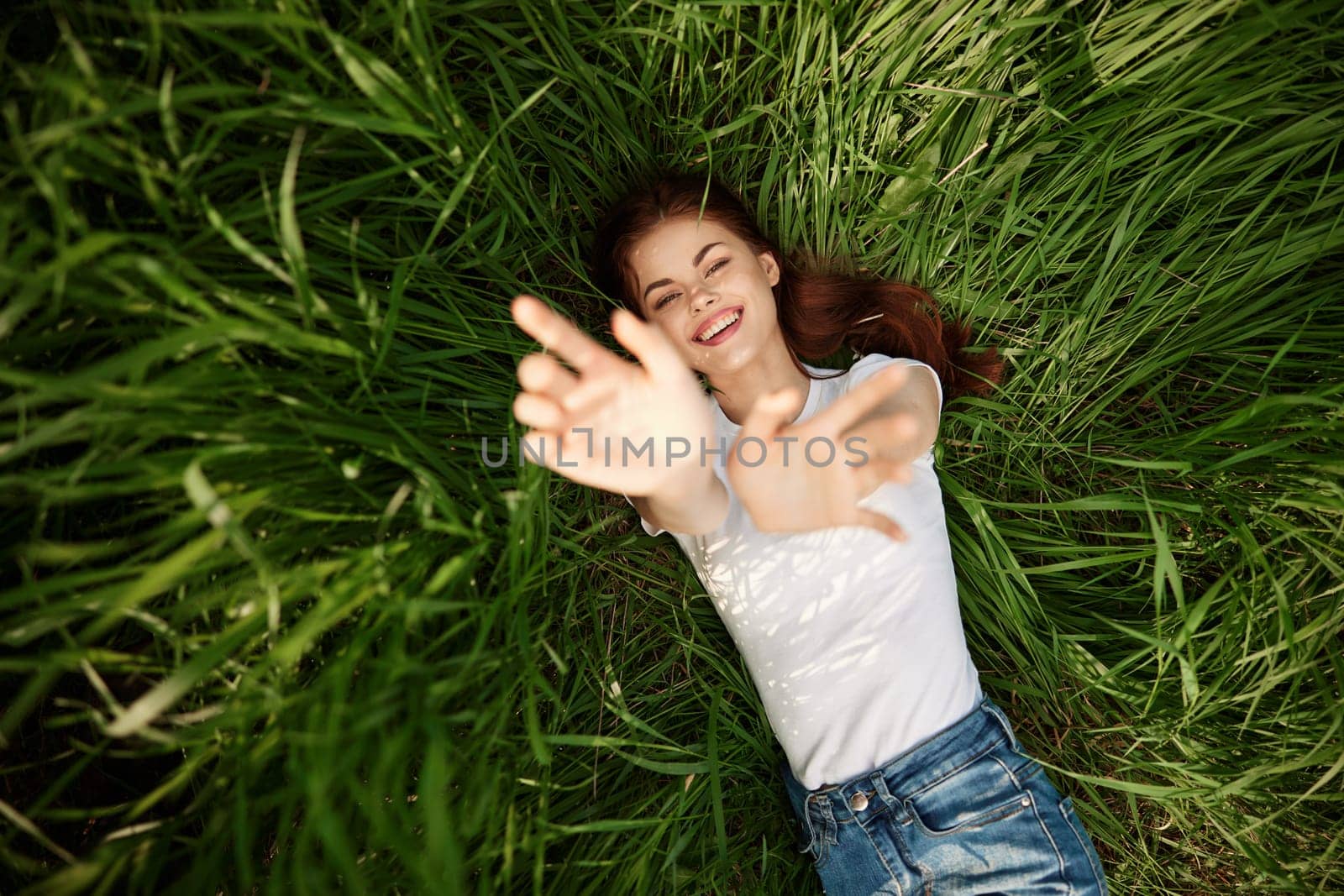 A happy girl in a white t-shirt stretches her arms up and lies on the green grass by Vichizh
