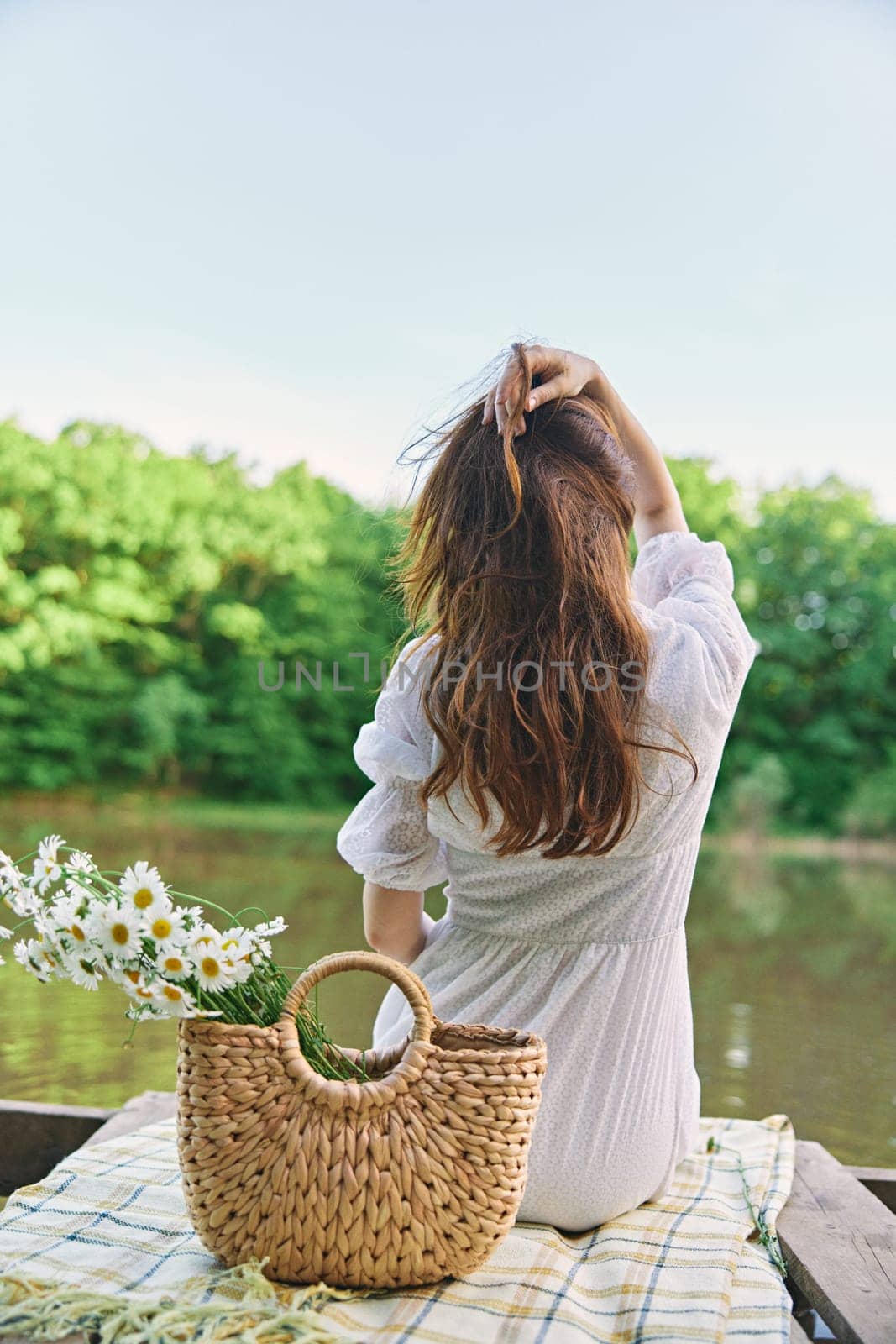 a woman in a light dress sits on the shore of the lake with her back to the camera and straightens her red hair. High quality photo