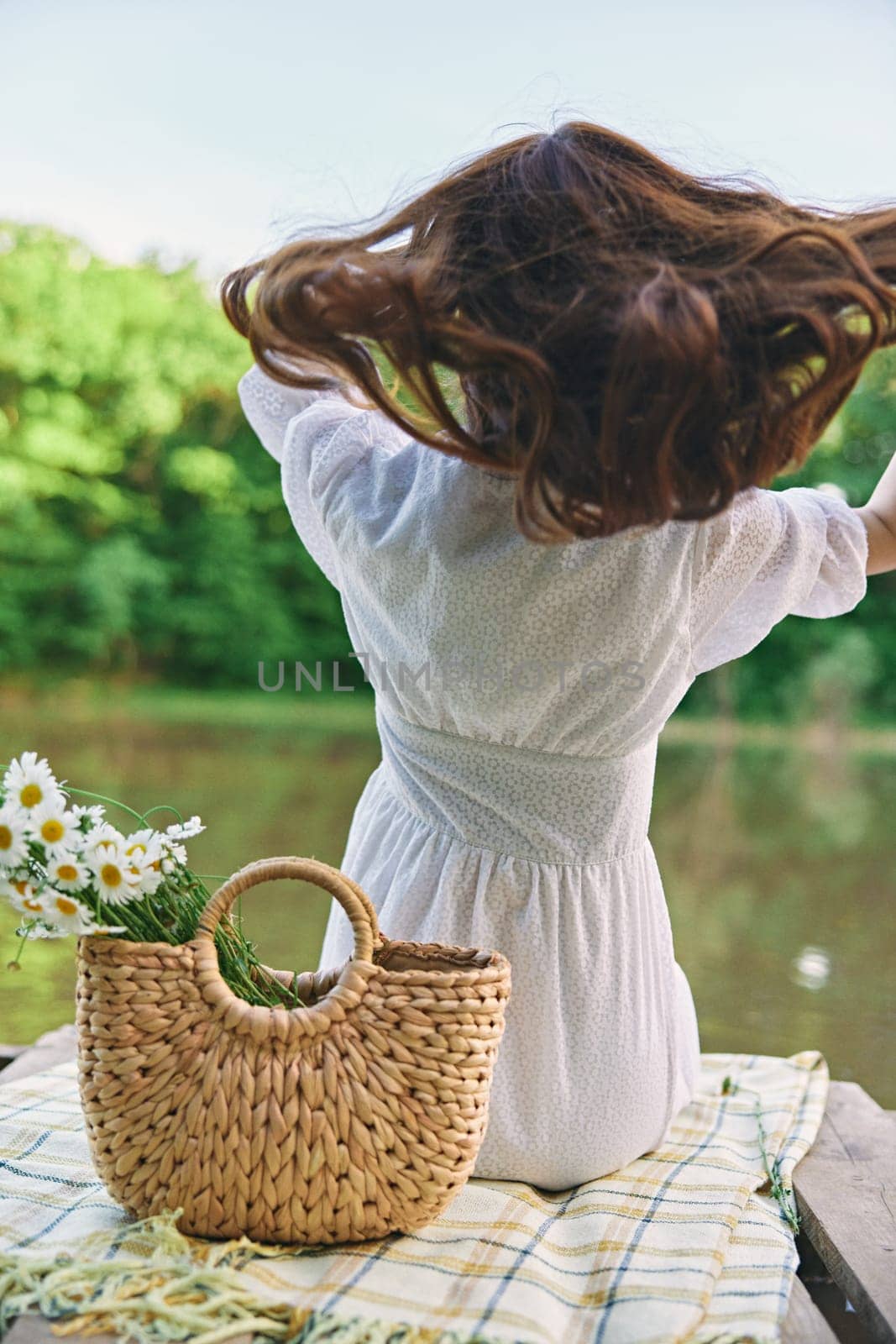 a woman in a light dress sits on the shore of the lake with her back to the camera and straightens her red hair by Vichizh