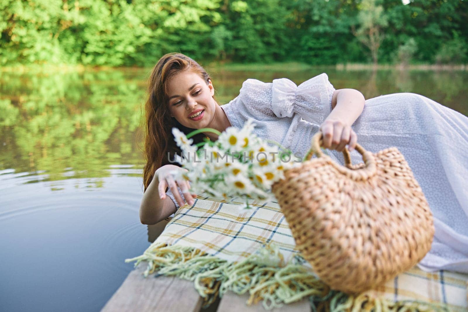 a lovely woman in a light dress lies on a pier by the lake on a sunny day with a basket of daisies in her hands. High quality photo