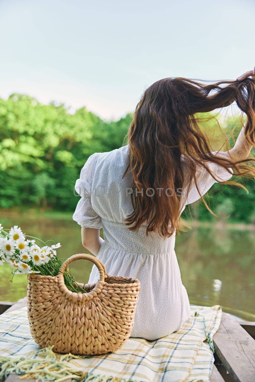 a woman in a light dress sits on the shore of the lake with her back to the camera and straightens her red hair. High quality photo