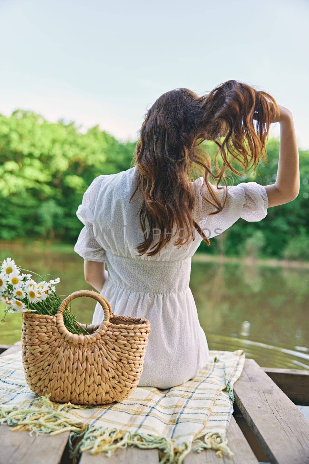 a woman in a light dress sits on the shore of the lake with her back to the camera and straightens her red hair by Vichizh