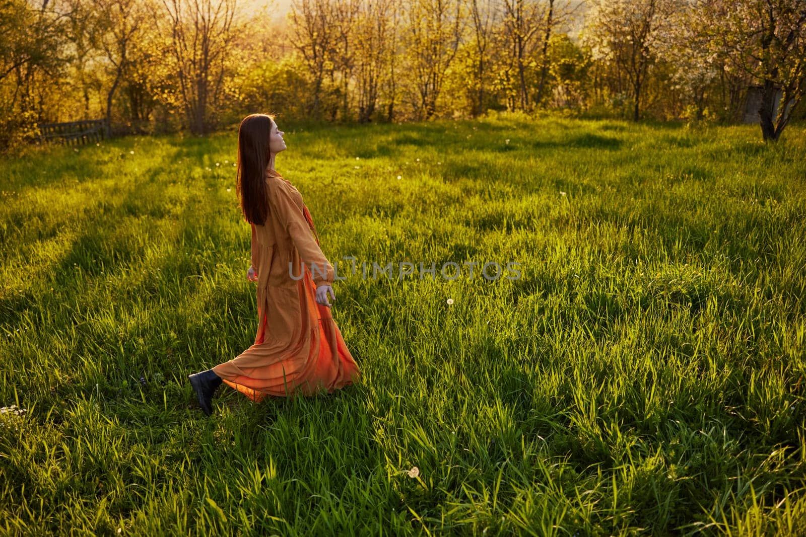 a joyful woman runs through a green field with her hands behind her back, enjoying a warm summer day and nature during the sunset. Horizontal photography in nature by Vichizh