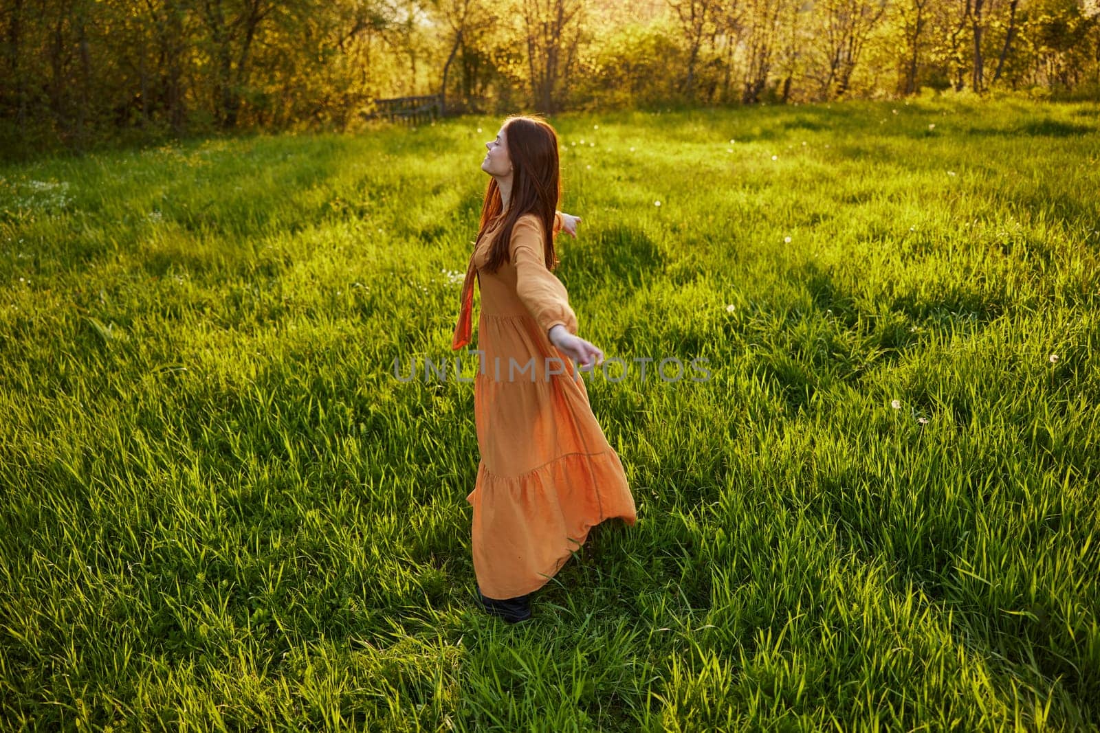 red-haired woman stands in a wide, green field during sunset in a long orange dress enjoying unity with nature and relaxation raising her arms to the sides while standing with her back to the camera by Vichizh