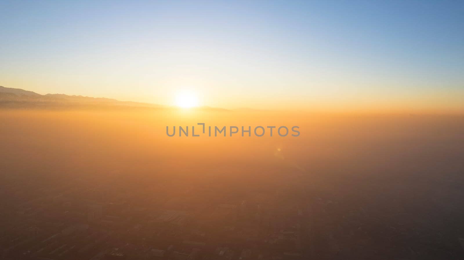 Epic gray smog is visible at sunset over the city. A bird's-eye view from a drone of houses, roads, cars and parks. White clouds and snowy mountains are illuminated by orange rays of the sun. Almaty