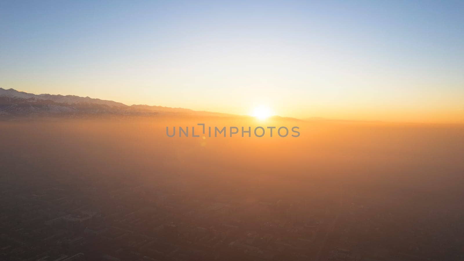 Epic gray smog is visible at sunset over the city. A bird's-eye view from a drone of houses, roads, cars and parks. White clouds and snowy mountains are illuminated by orange rays of the sun. Almaty