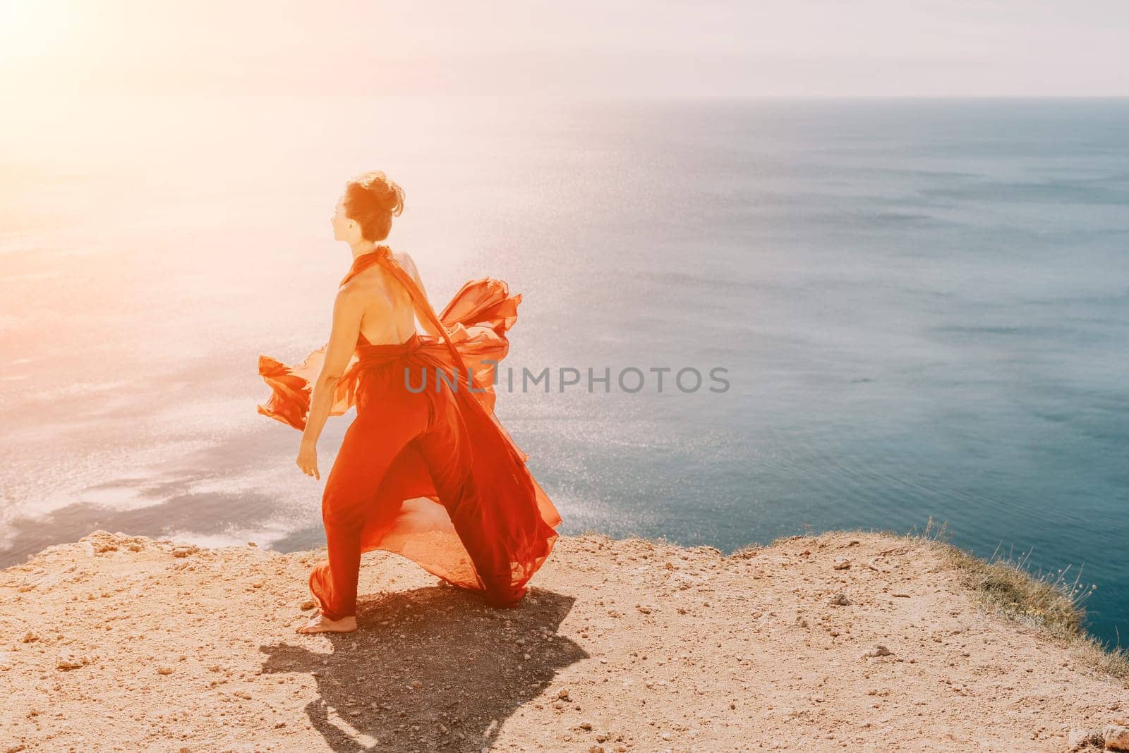 Side view a Young beautiful sensual woman in a red long dress posing on a rock high above the sea during sunrise. Girl on the nature on blue sky background. Fashion photo.