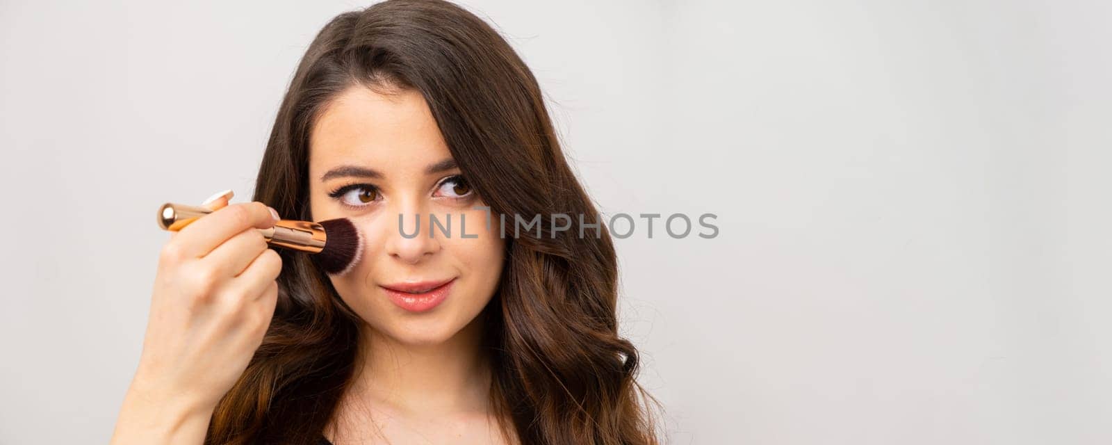 Brunette beautician applies powder on face. Young woman uses big brush to do makeup on grey background with copy space
