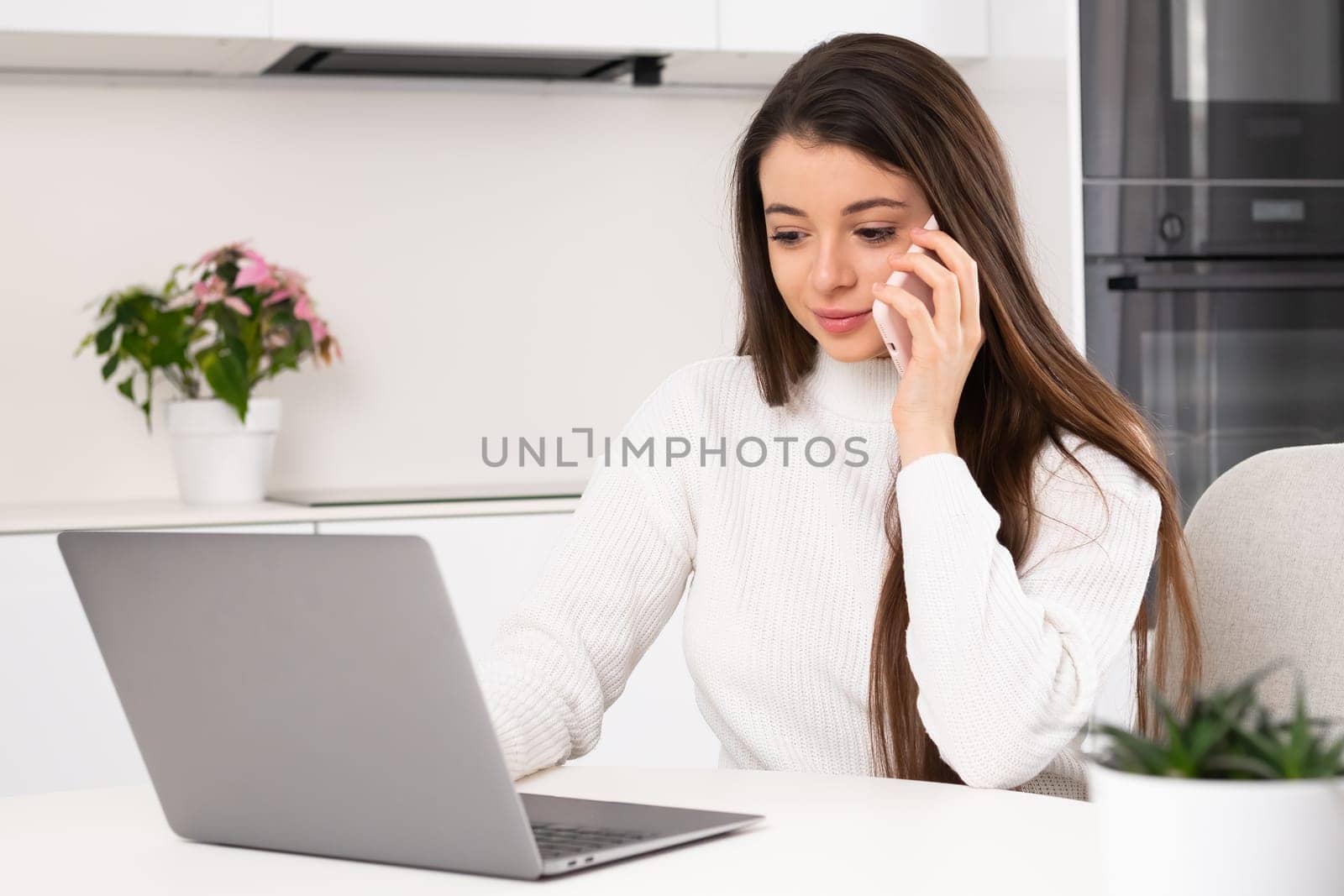 Woman has a call using smartphone while working with laptop at home.