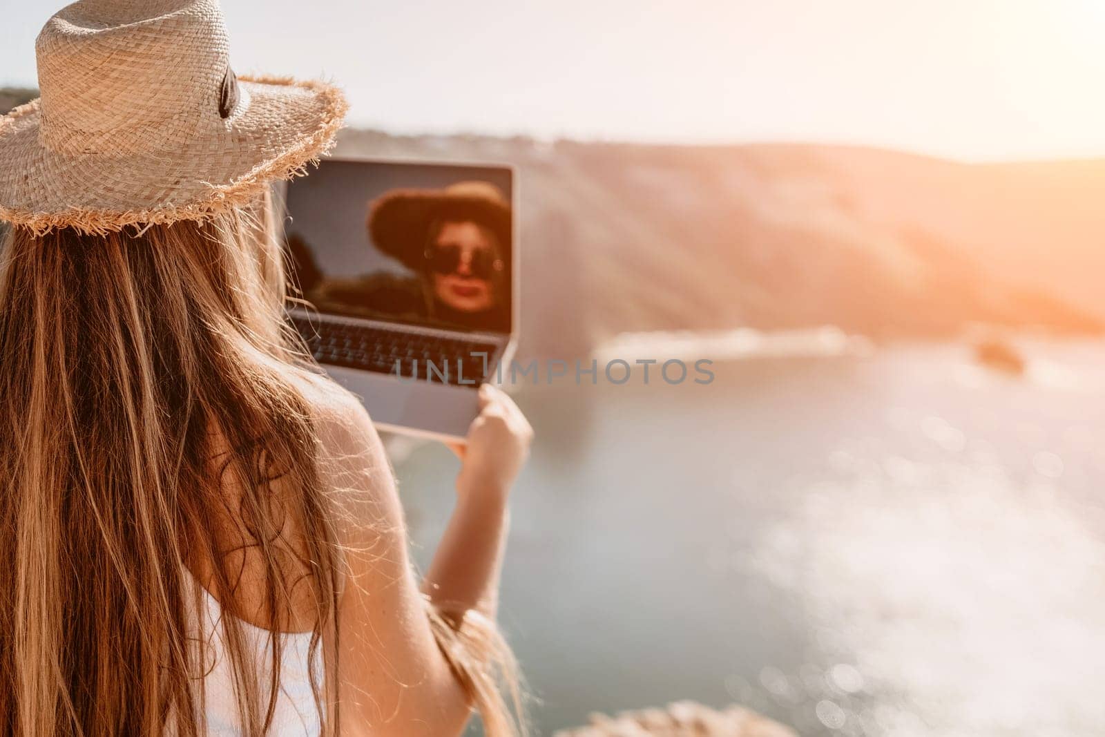 Successful business woman in yellow hat working on laptop by the sea. Pretty lady typing on computer at summer day outdoors. Freelance, travel and holidays concept.