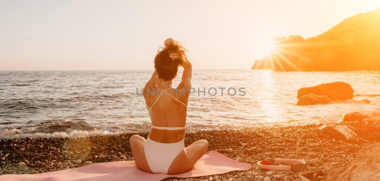 Young woman in swimsuit with long hair practicing stretching outdoors on yoga mat by the sea on a sunny day. Women's yoga fitness pilates routine. Healthy lifestyle, harmony and meditation concept.