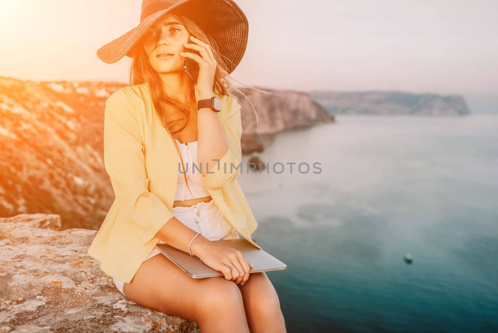 Successful business woman in yellow hat working on laptop by the sea. Pretty lady typing on computer at summer day outdoors. Freelance, travel and holidays concept.