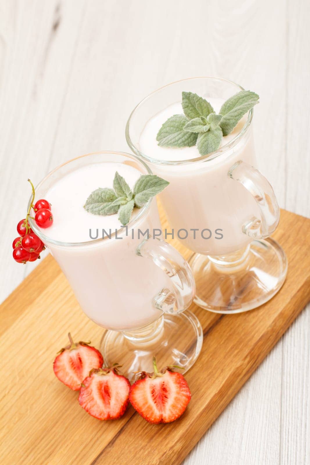 Two glasses of delicious strawberry yogurt with mint leaves and fresh strawberries on wooden cutting board.