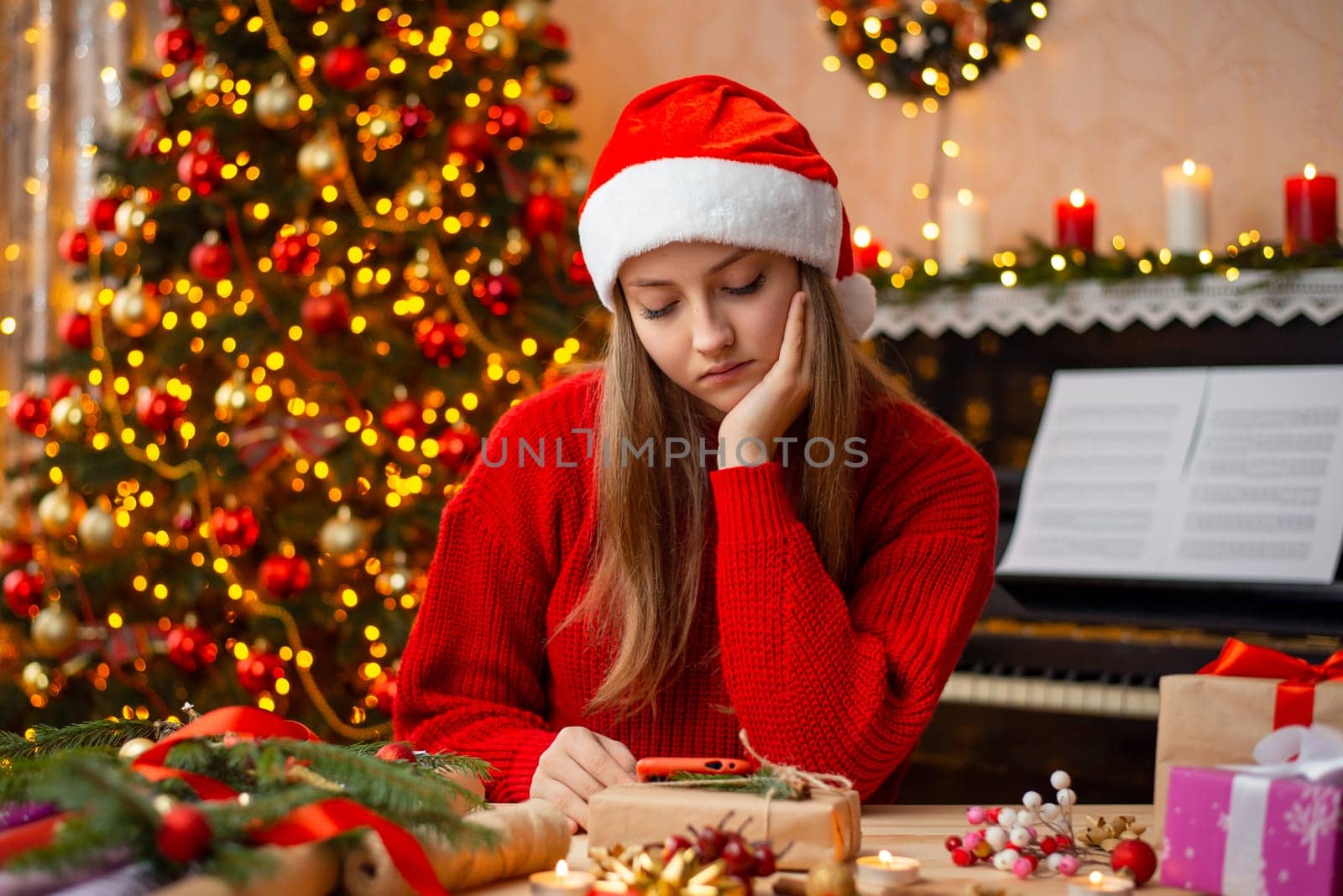 Sad young girl in red sweater and santa hat sits at the table and watches something on smartphone