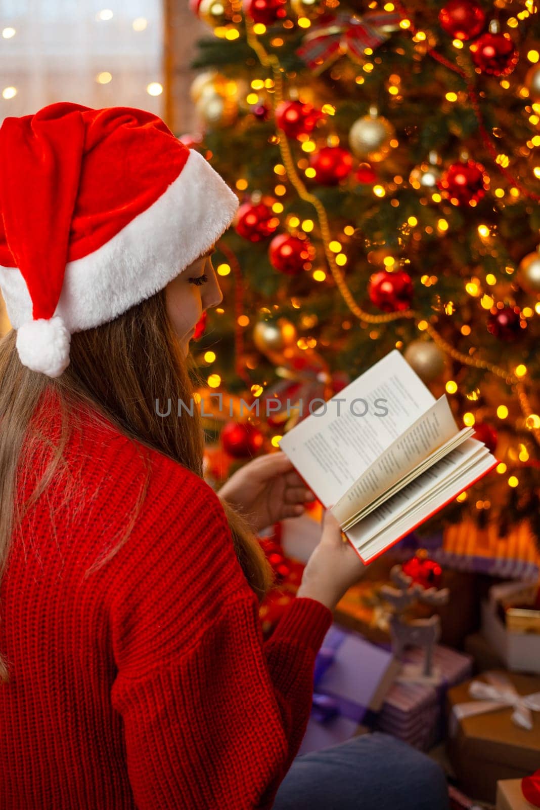 Vertical photo of young woman sitting near decorated Christmas tree and reading a book