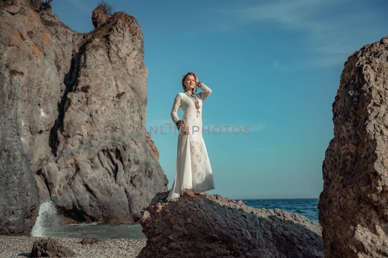 Woman white dress sea stones rocks.Middle-aged woman looks good with blond hair, boho style in a white long dress on beach jewelry around her neck and arms