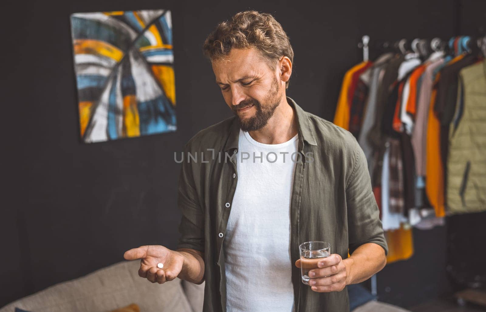 Man holding pill in one hand and glass of water in the other, isolated at home. Concepts of healthcare, wellness, and staying home for quarantine or isolation. The man is indoors and focused on taking his medication, while the glass of water provides a sense of refreshment and hydration. High quality photo