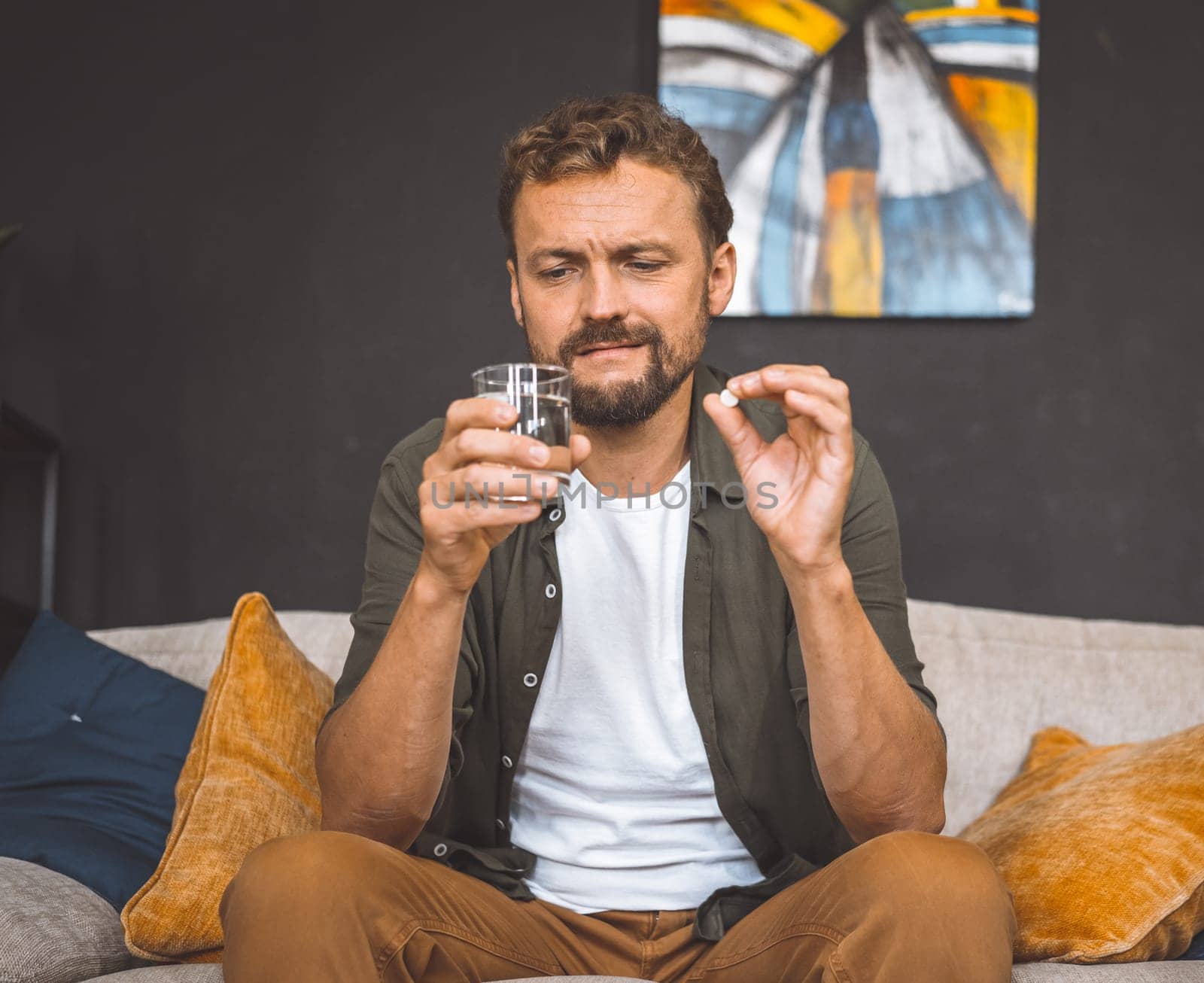 Caucasian man sit on sofa home, holding pill in one hand and glass of water in other. He focused on taking medicine, as he is dealing with illness or health condition that requires medication. High quality photo