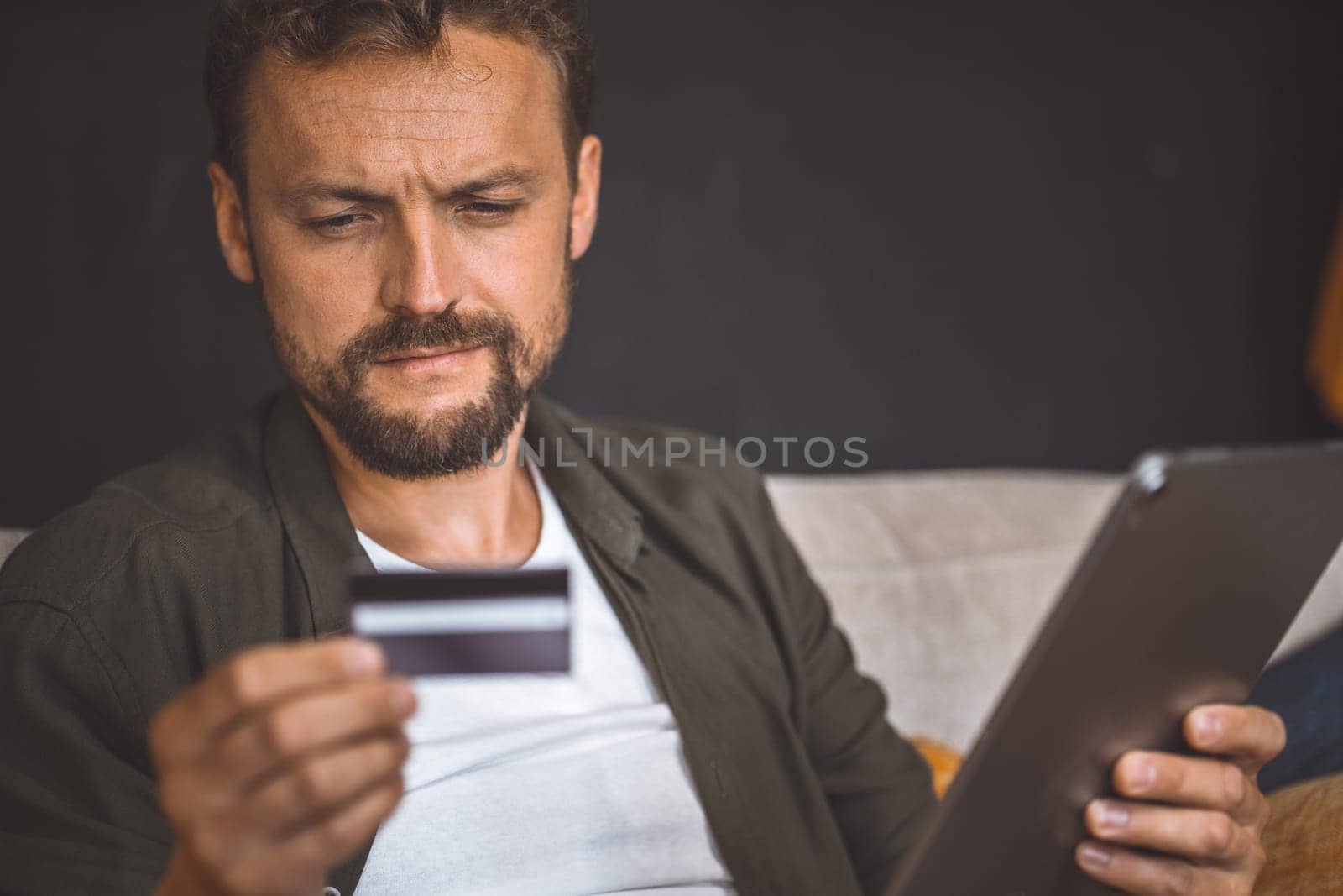 Man is seen sitting home and using his tablet PC for online shopping. He holding credit card in one hand and the tablet PC in the other, and making payment on a website. Ease of online shopping. High quality photo