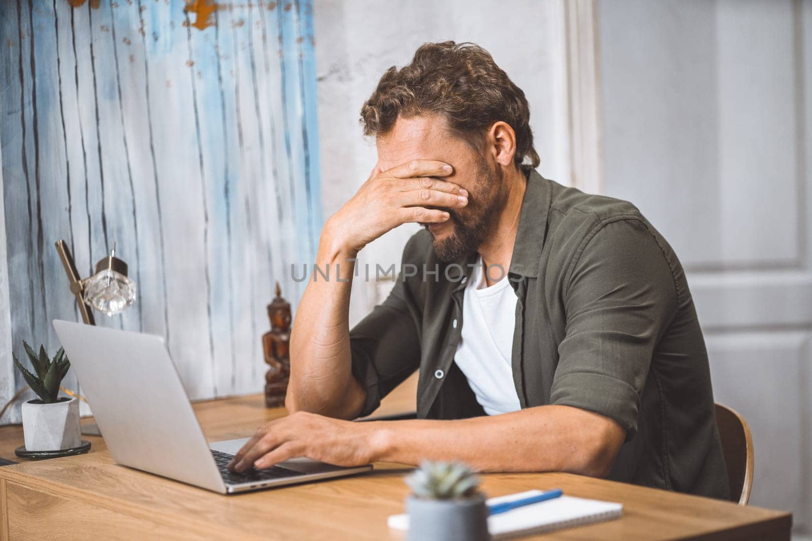 Man clearly overwhelmed and stressed out at work. His head in his hands, covering his face with palm, appearing to be in state of deep depression. Computer in front of him serves as a reminder of the pressures and deadlines he is facing, and the overall image portrays a feeling of burnout and exhaustion. by LipikStockMedia