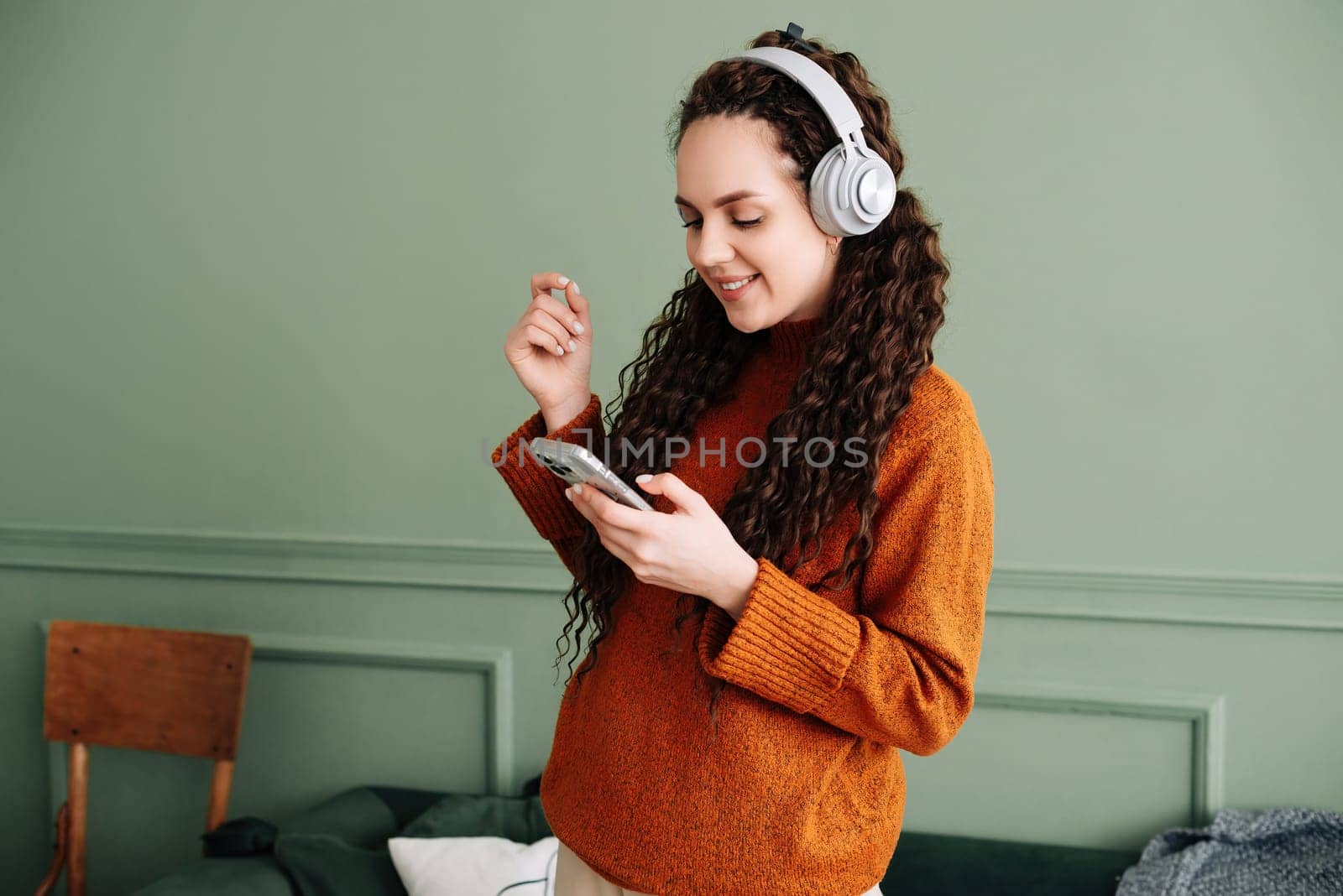 Happy pretty young woman wearing headphones dancing at home. Cheerful girl listening to music on mobile phone, singing songs feeling relaxed standing in modern living room interior