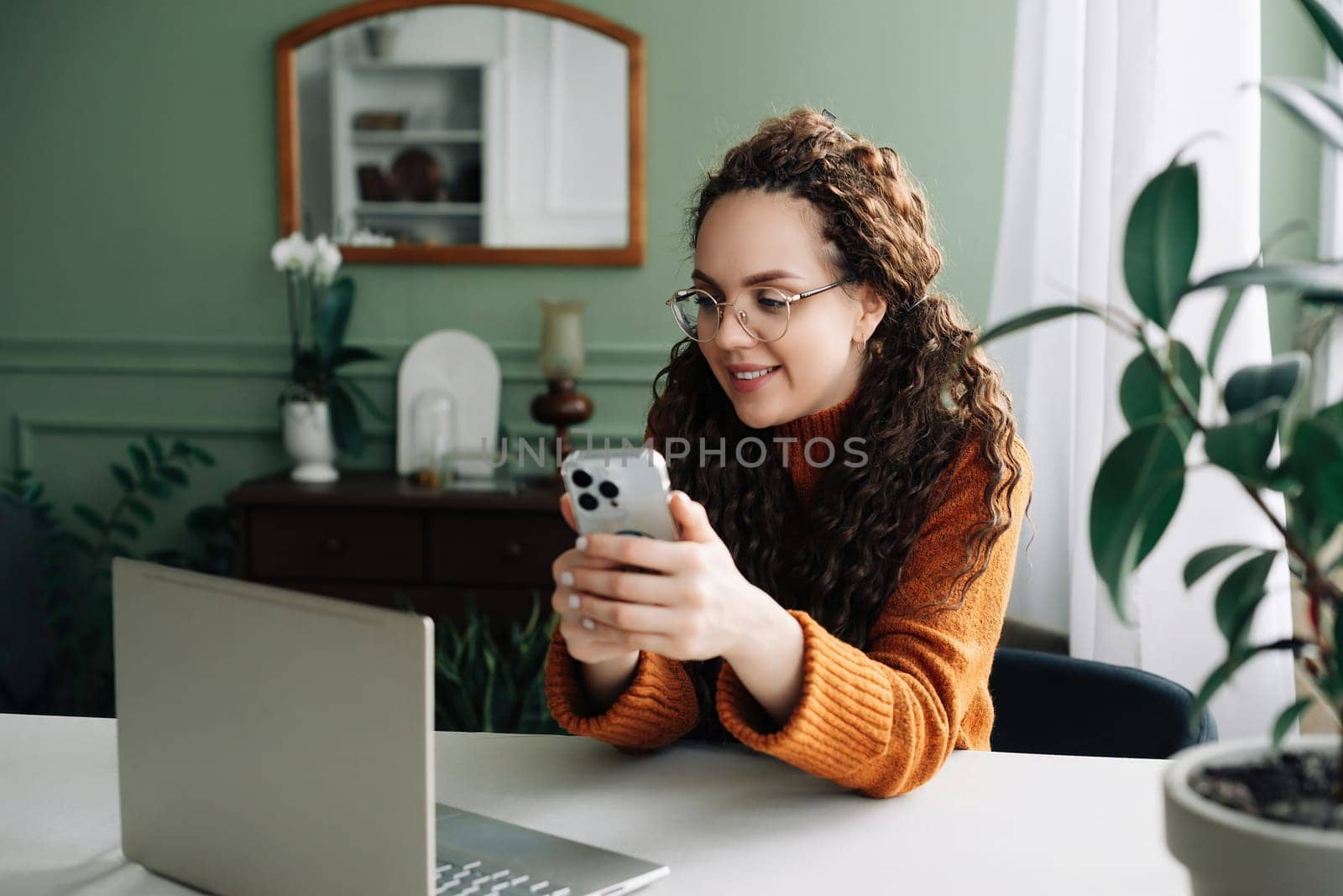 Young happy smiling pretty woman sitting at table holding smartphone using cellphone modern technology, looking at mobile phone while remote working or learning, texting messages at home