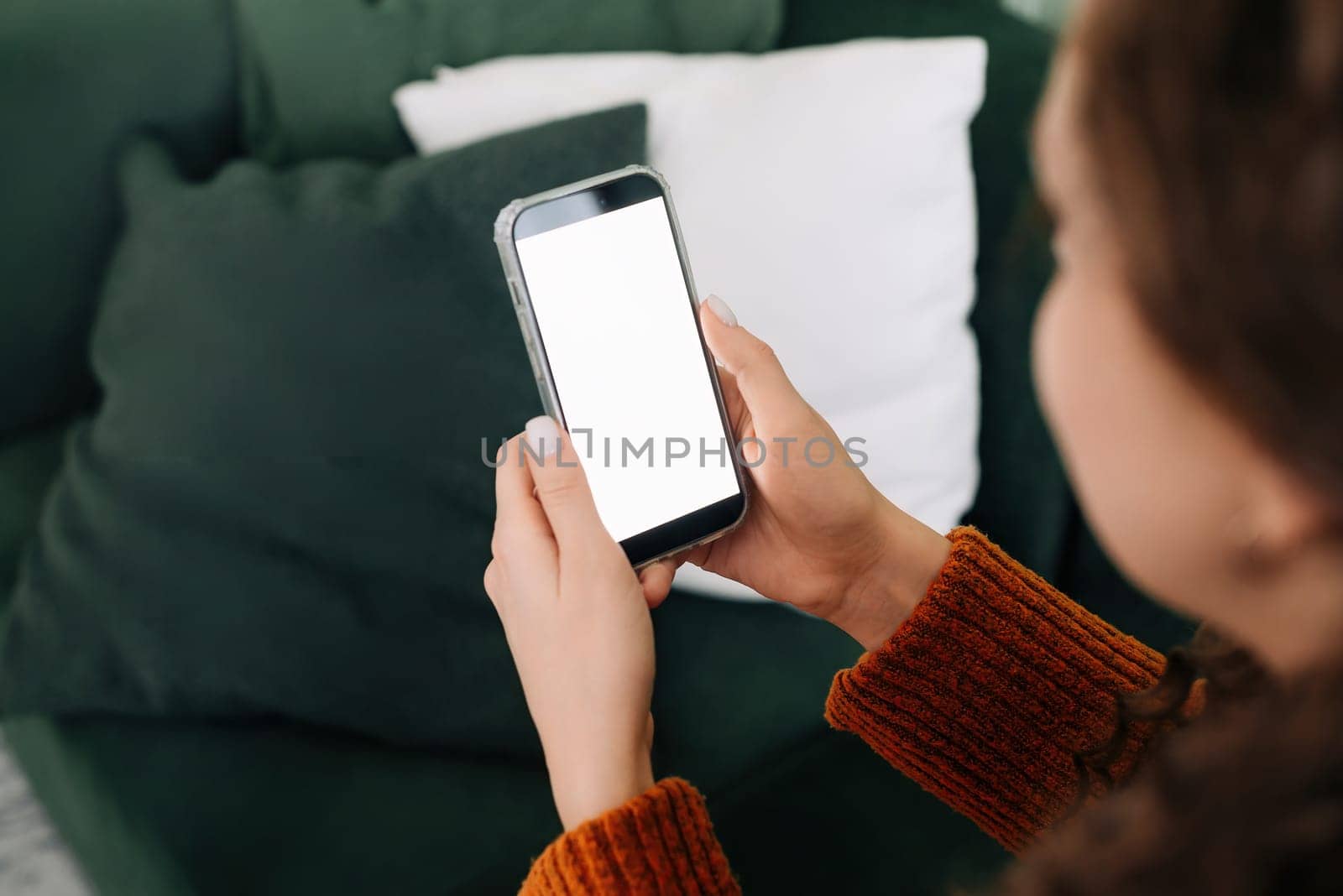 Over shoulder view of woman holding smartphone with white mock up cellular screen in hands using mobile apps on cell phone relaxing on sofa at home. Cellphone display mock up for advertising concept