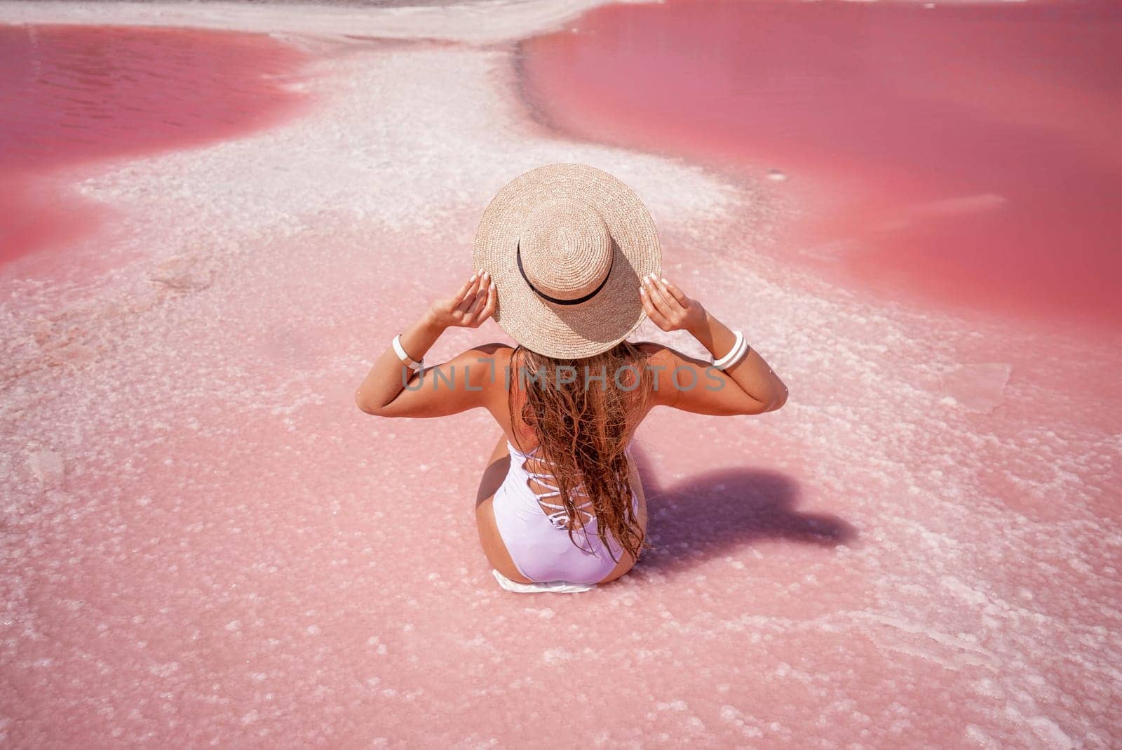A woman traveler looks at an amazing pink salt lake. He sits with his back in a bathing suit and holds his hat in his hands. Wanderlust travel concept by Matiunina