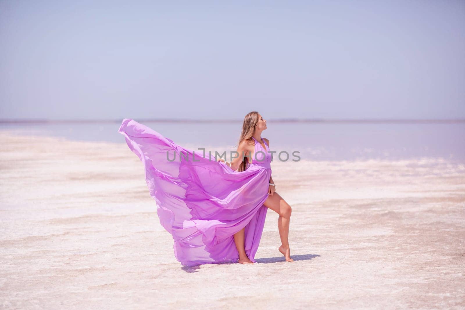 A woman in a pink dress enjoys the scenic view of a pink salt lake as she walks along the white, salty shore, creating a lasting memory. by Matiunina